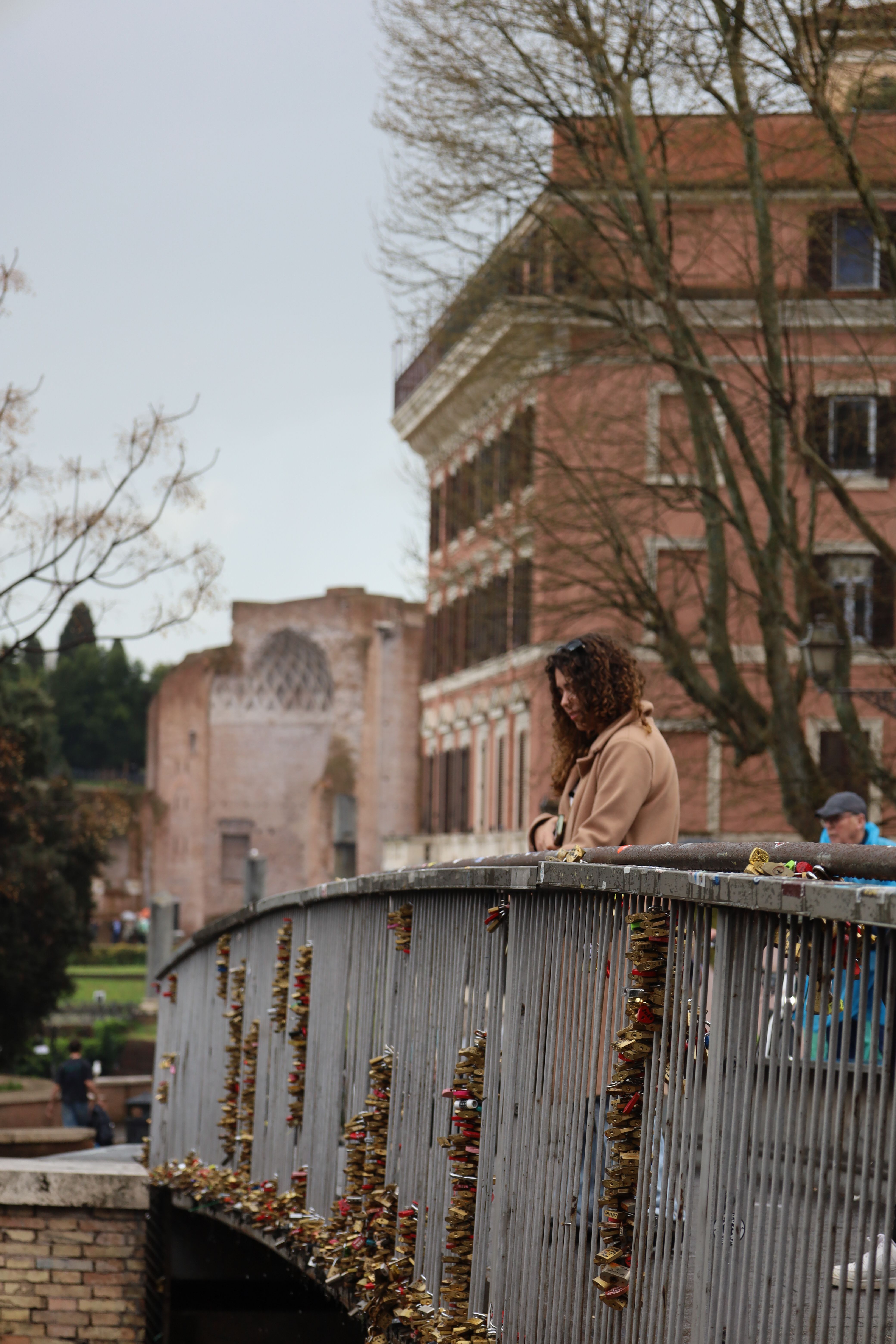 A woman contemplatively gazes from a bridge adorned with locks in Rome, capturing the essence of urban serenity.