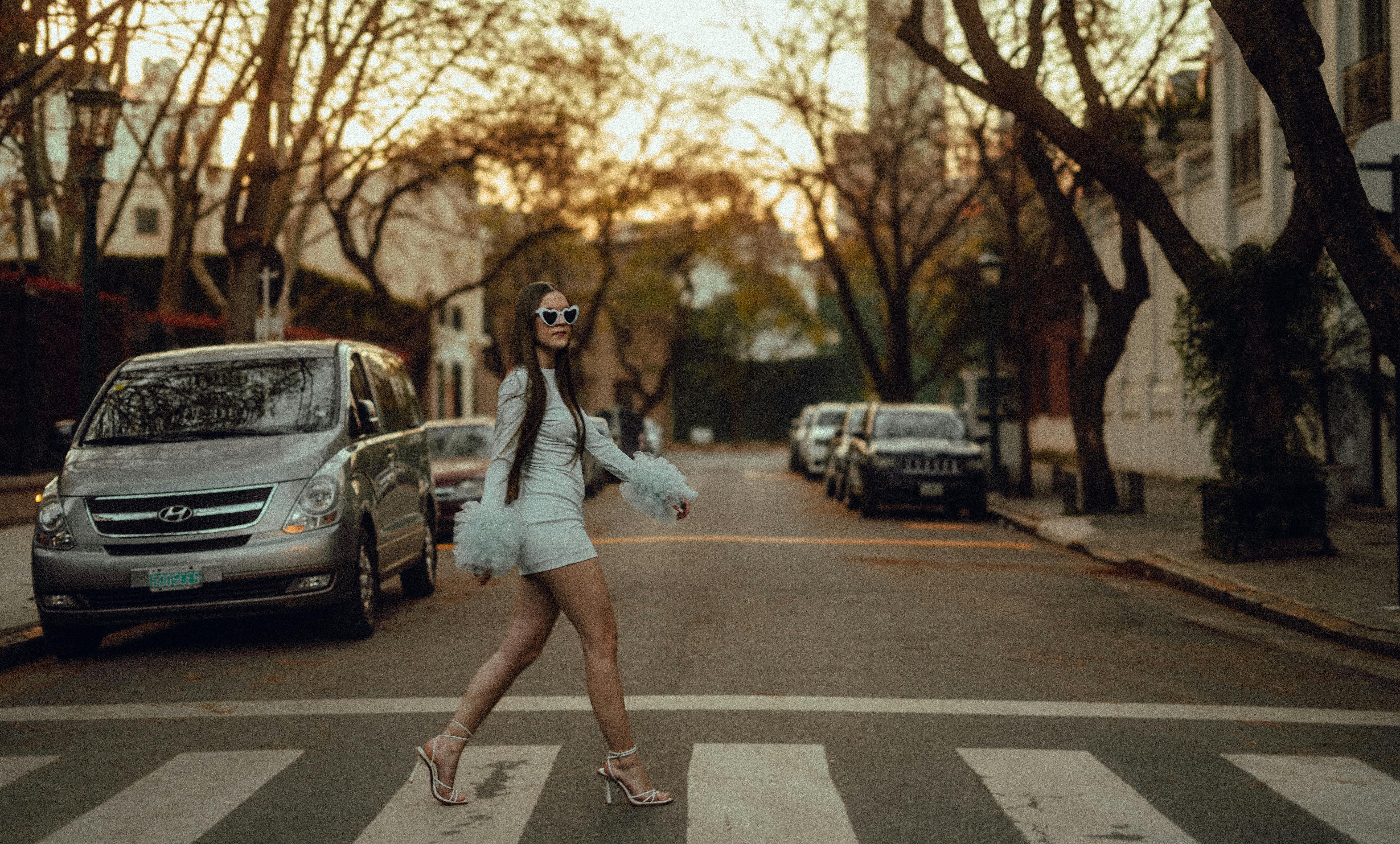 무료 세련된 옷차림의 우아한 여성이 석양 아래 도시 거리를 건너며 자신감을 뽐내고 있다. 스톡 사진