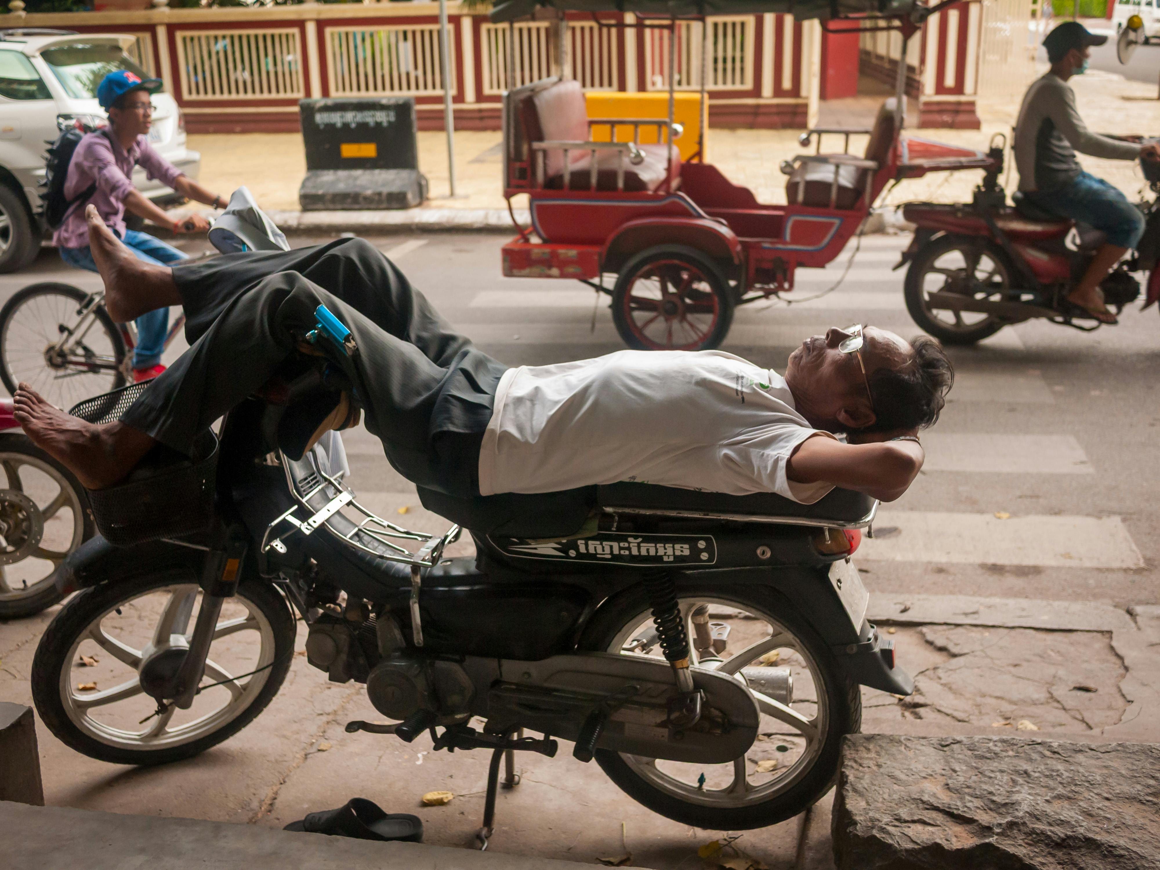 免费 在柬埔寨金边熙熙攘攘的街道上，一名男子悠闲地斜倚在一辆停着的摩托车上。 素材图片