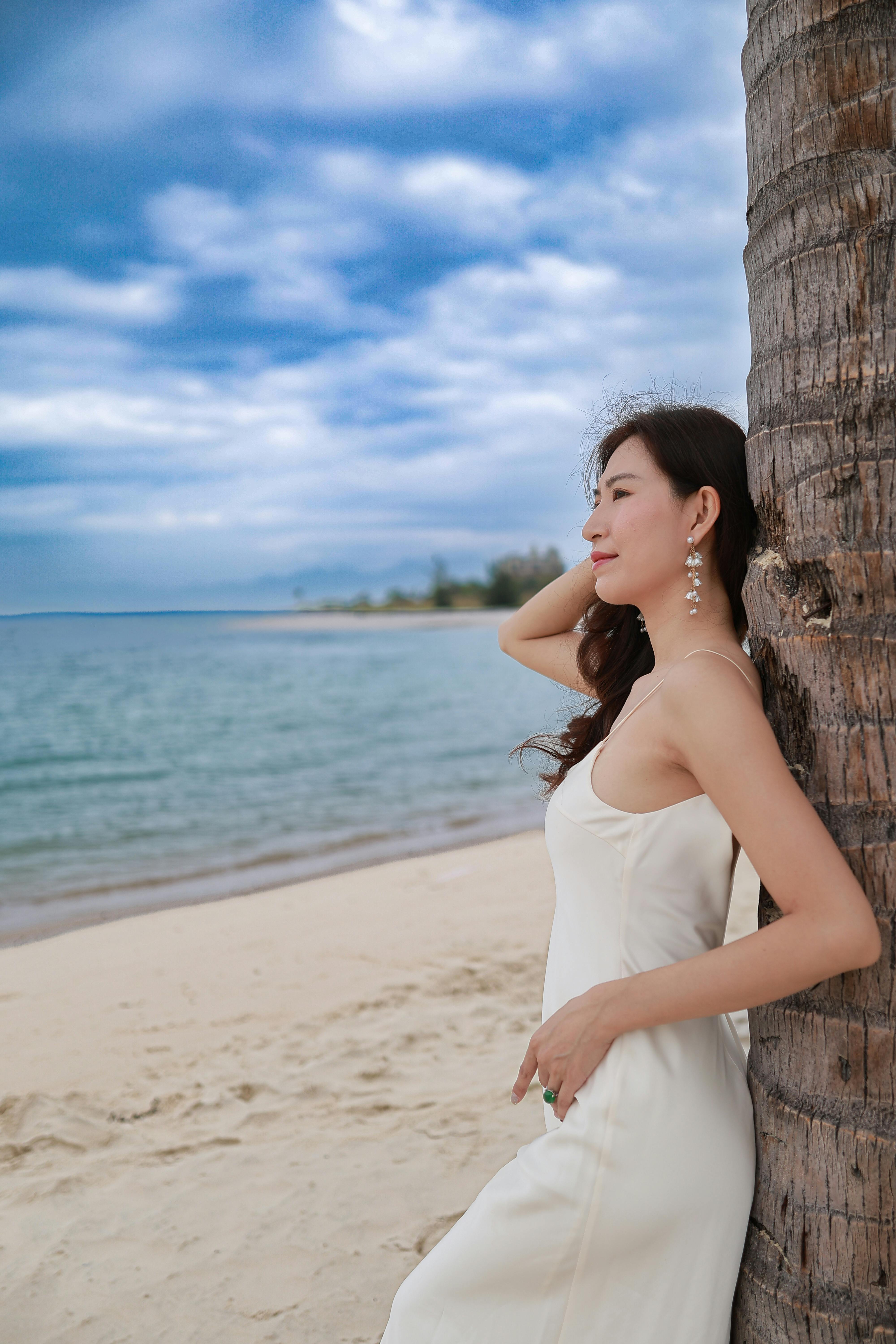 A serene woman in a white dress leans against a palm tree on Nha Trang Beach, Vietnam.