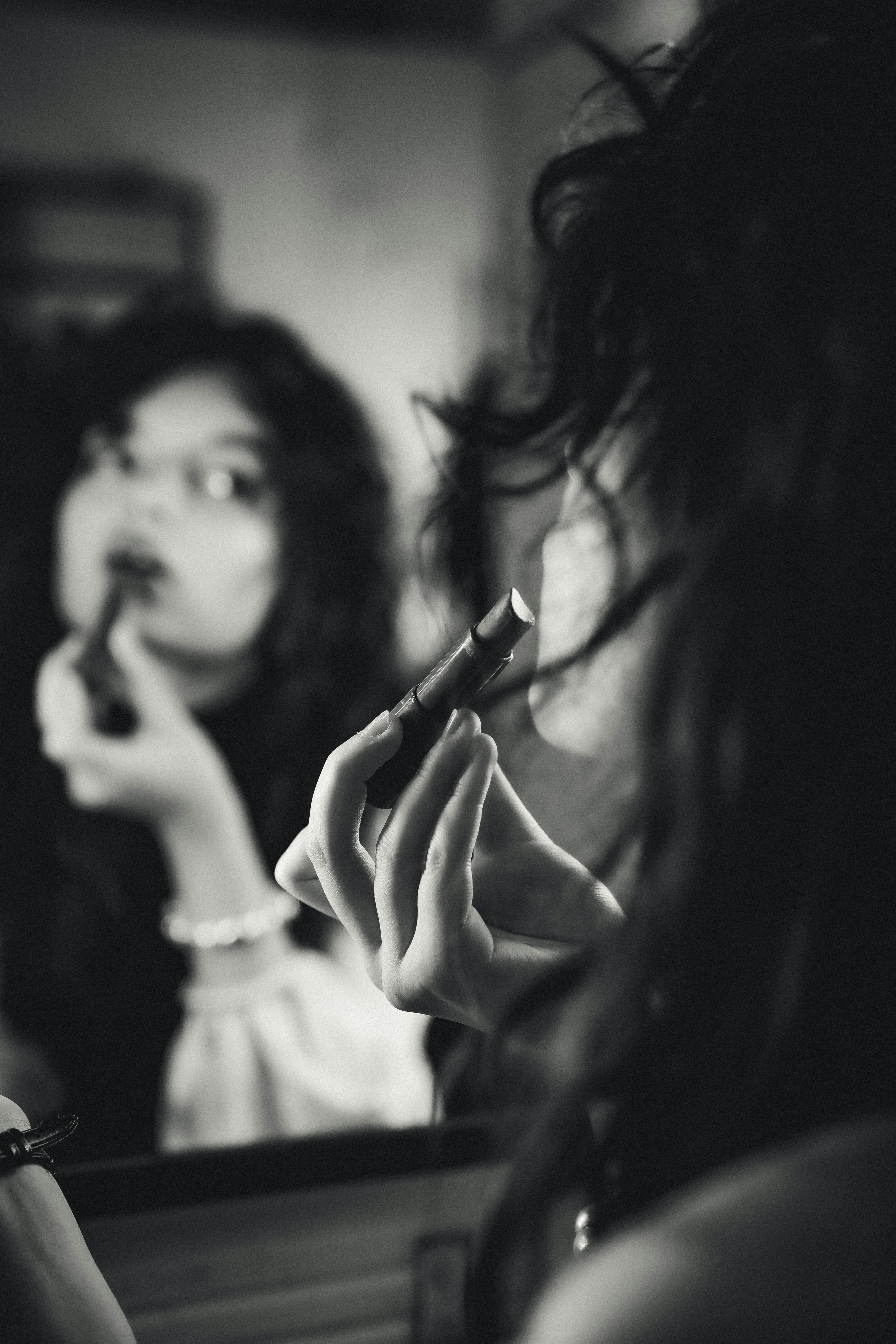 Безкоштовне Монохромне зображення жінки, яка наносить темну помаду перед дзеркалом, що фіксує момент особистого роздуму. стокове фото