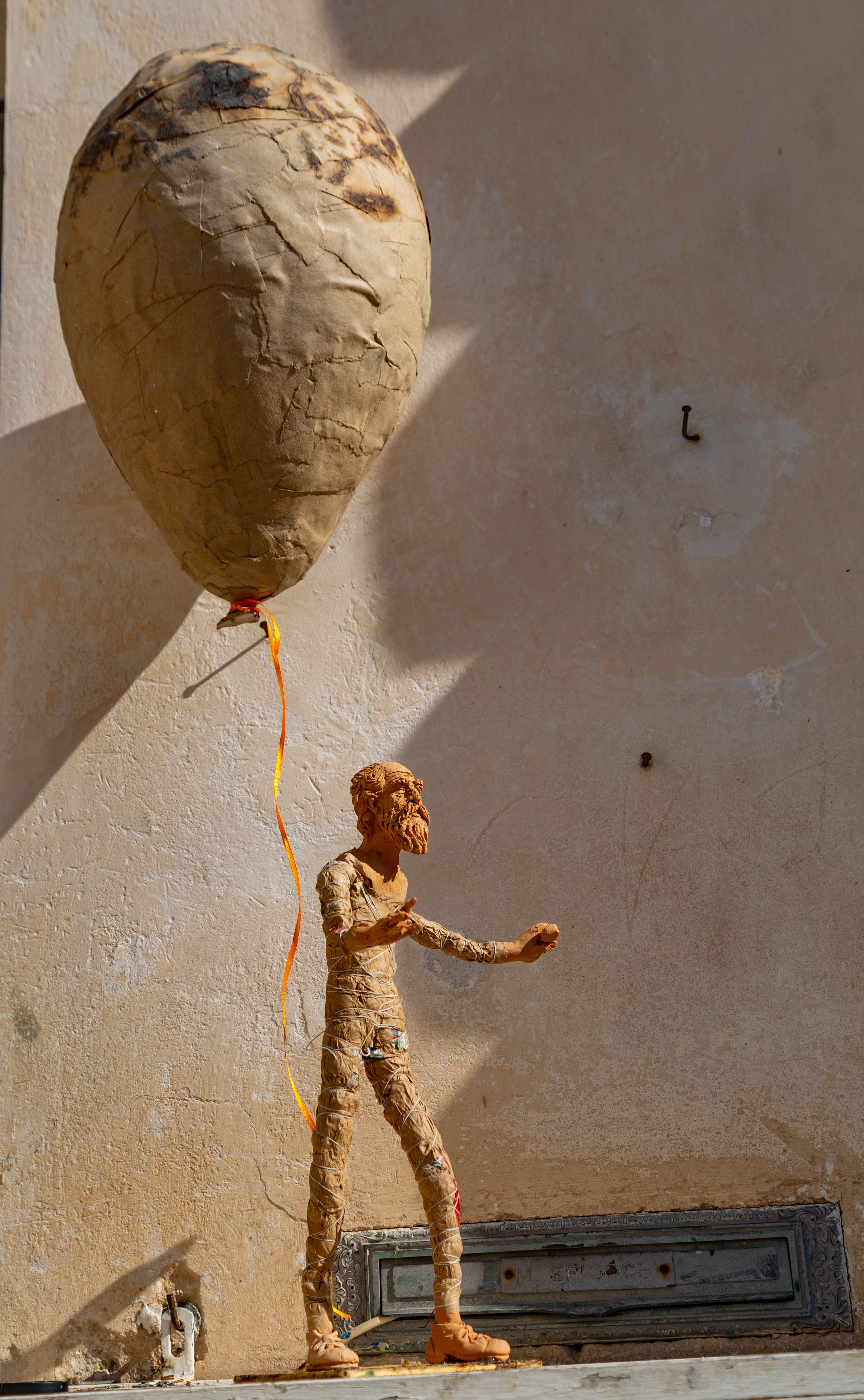 Paper mache figure with large balloon photographed against a wall in Bari, Italy, highlighting texture and artistry.
