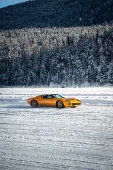Vintage yellow car driving on a snow-covered road with forest and mountain background.