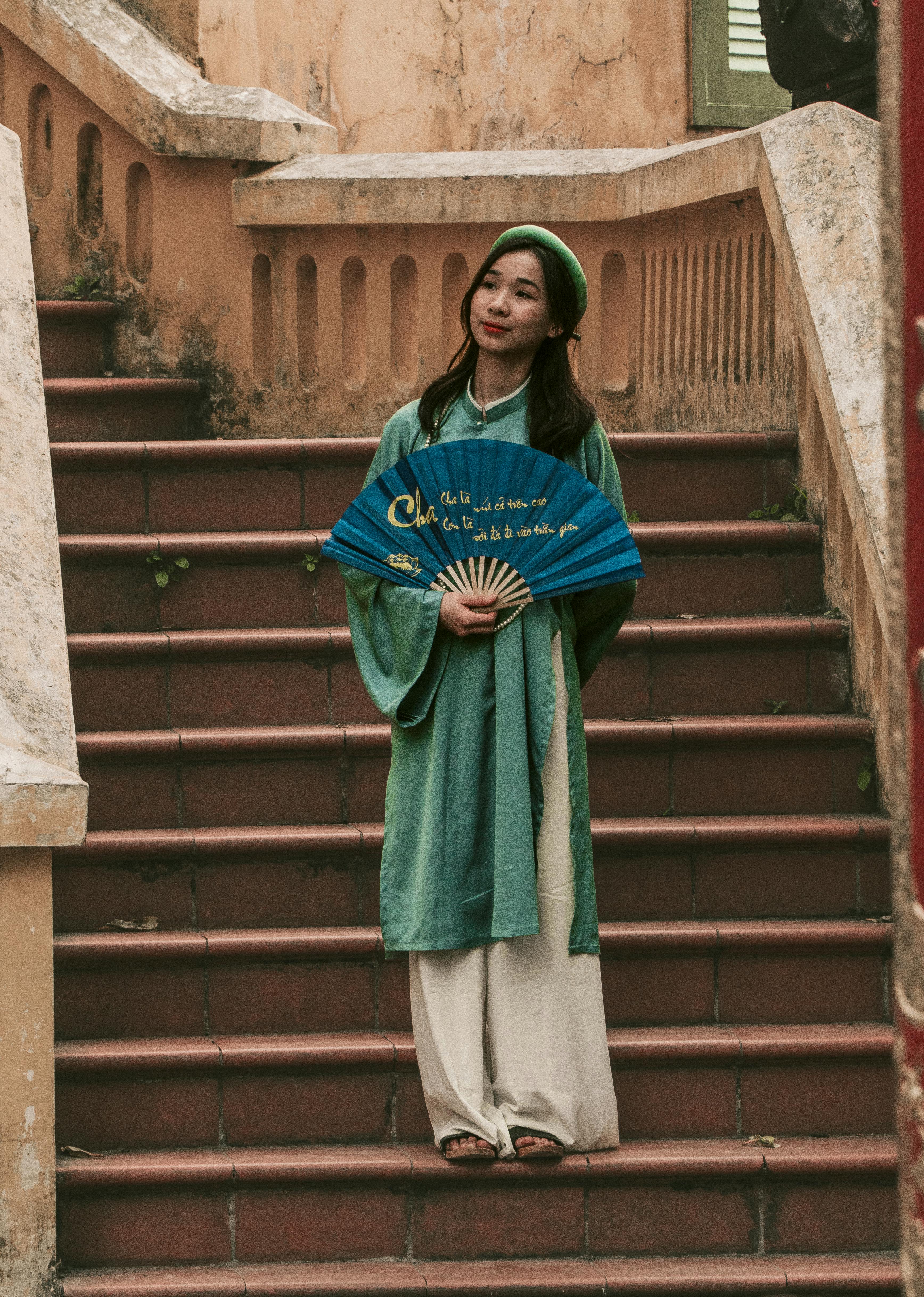 免费 一位身着越南传统服饰的年轻女子站在台阶上，手里拿着一把扇子。 素材图片