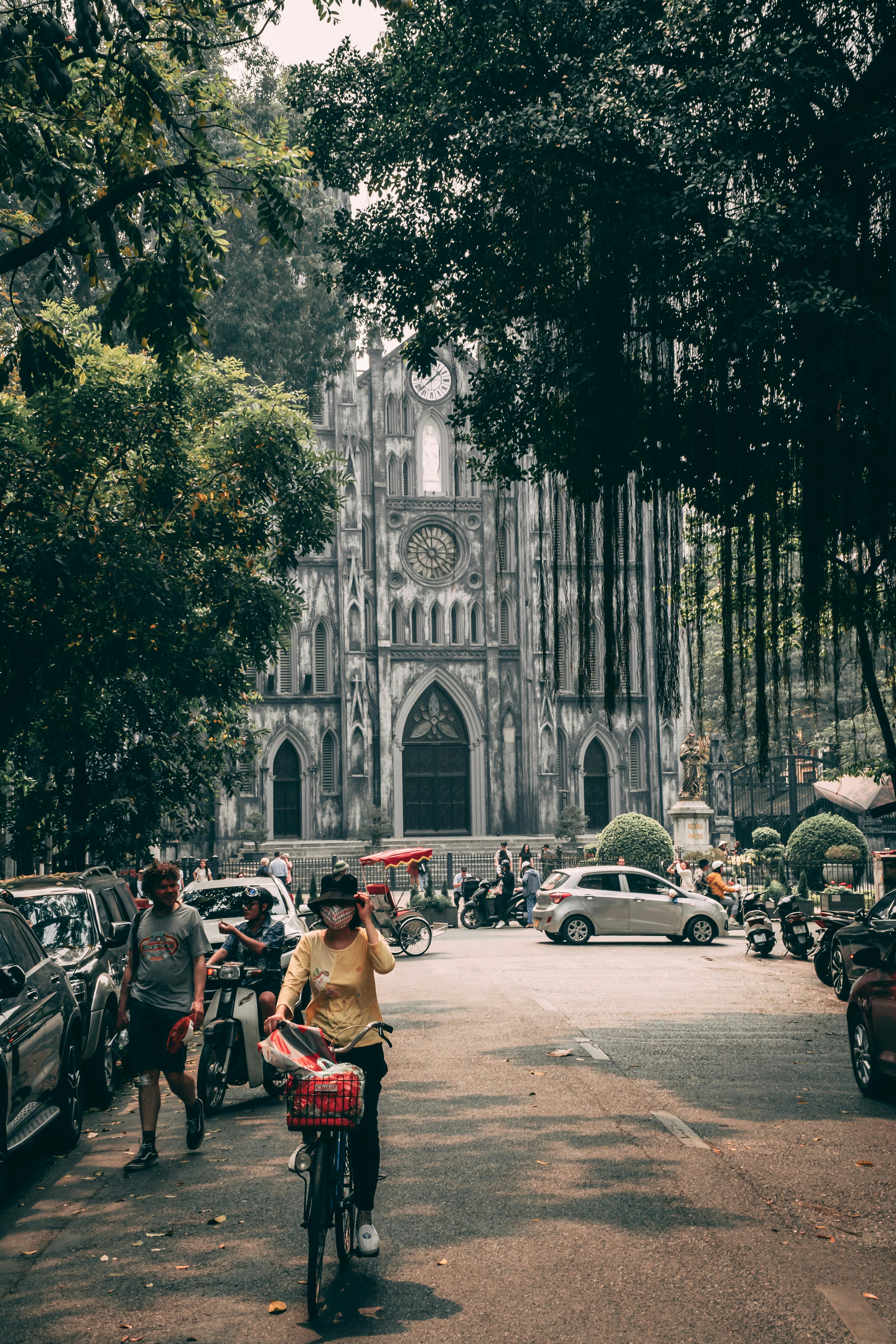 免费 自行车和行人从一座历史悠久的大教堂旁经过，教堂周围绿树成荫，停满了汽车。 素材图片