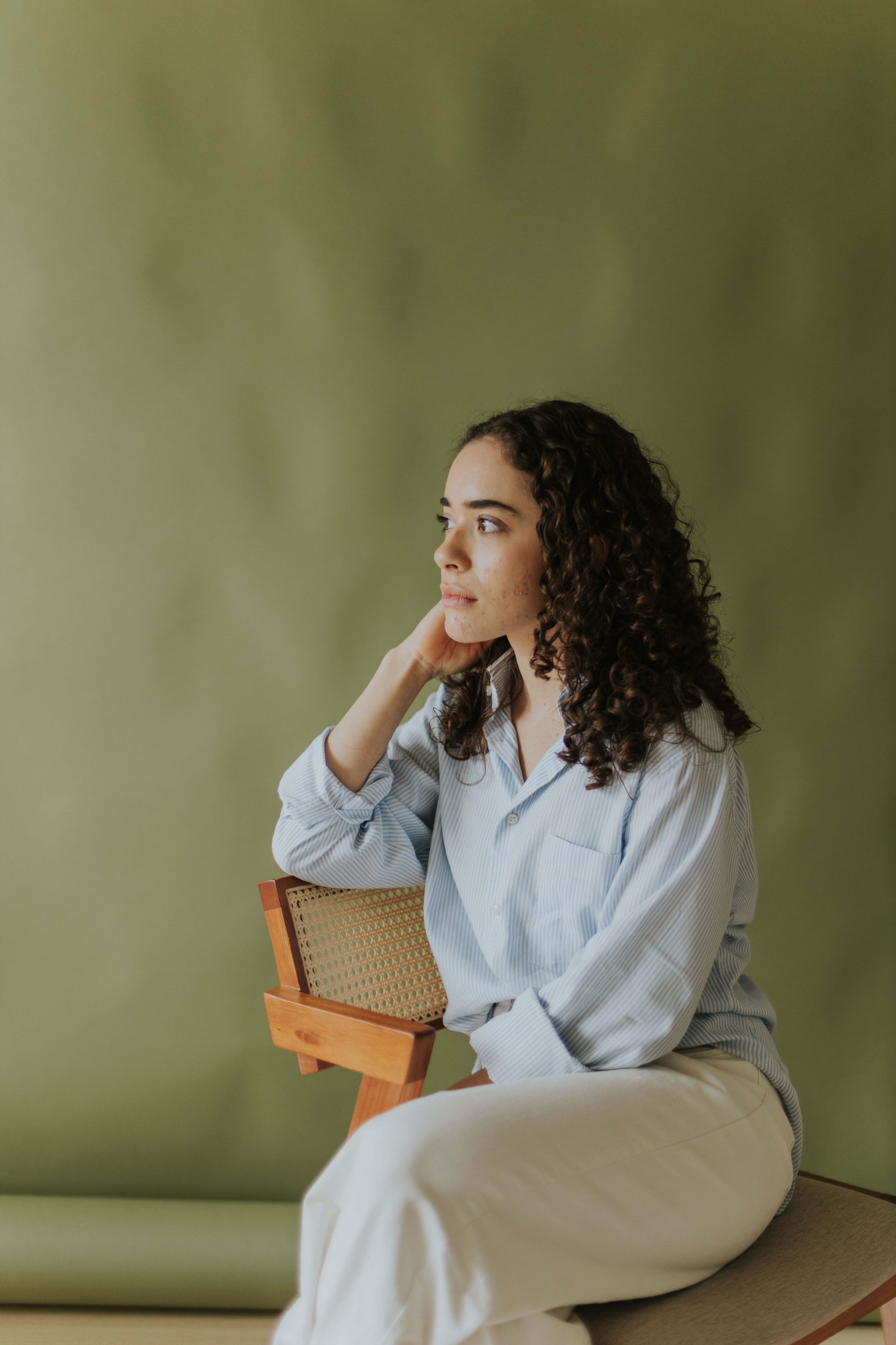 A contemplative woman sitting on a chair indoors against a green background, wearing casual clothing.