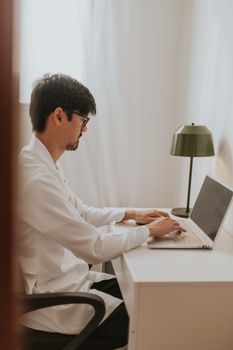 A man in a white shirt works on a laptop in a cozy home office setting.