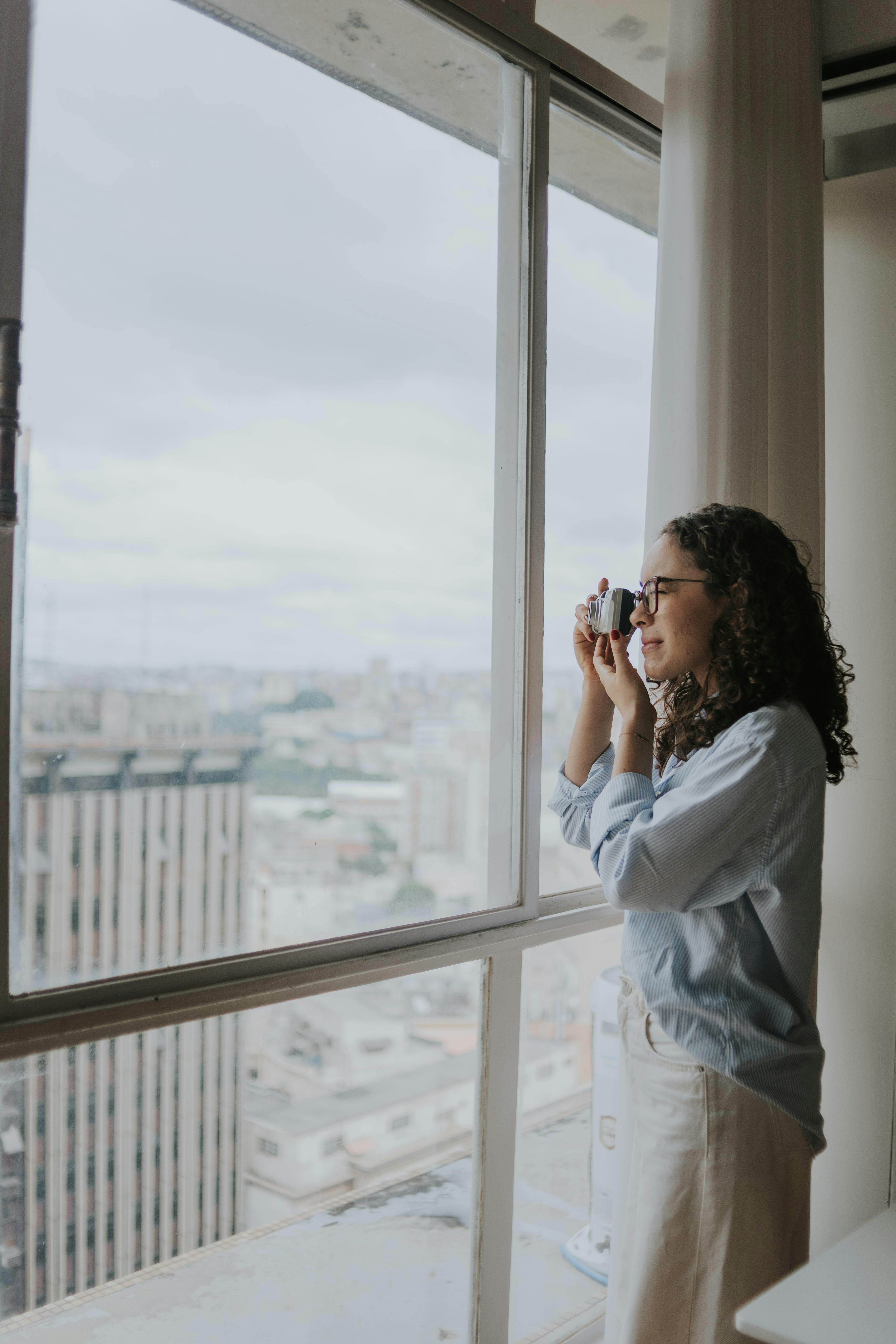 ingyenes Oldalnézetből egy nő kamerával rögzíti a város látképét egy ablakon keresztül egy borús napon. Stockfotó