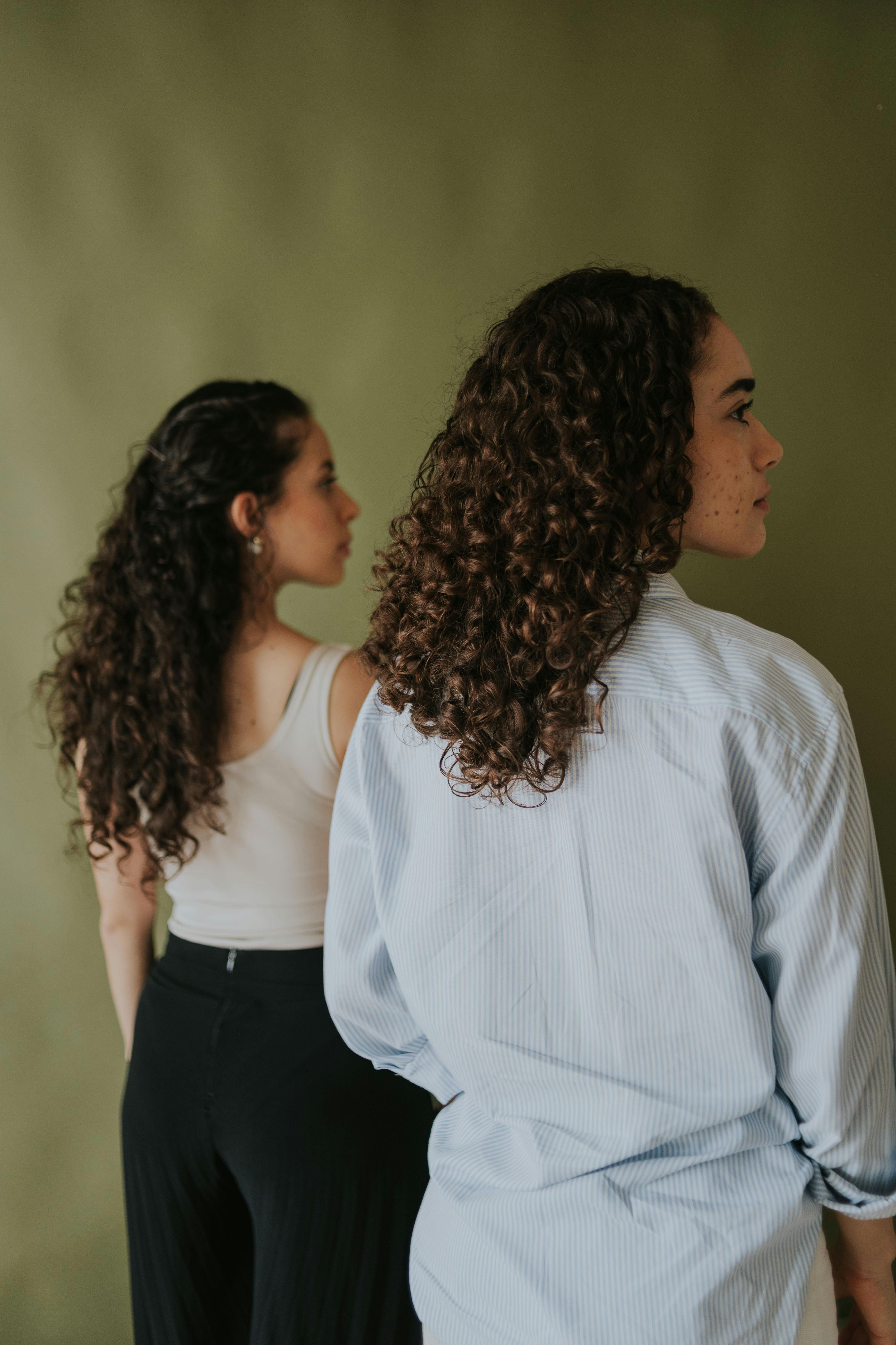 Miễn phí Hai người phụ nữ tóc xoăn đứng quay lưng vào nhau trước một phông nền đơn giản, trang phục thường ngày. Ảnh lưu trữ