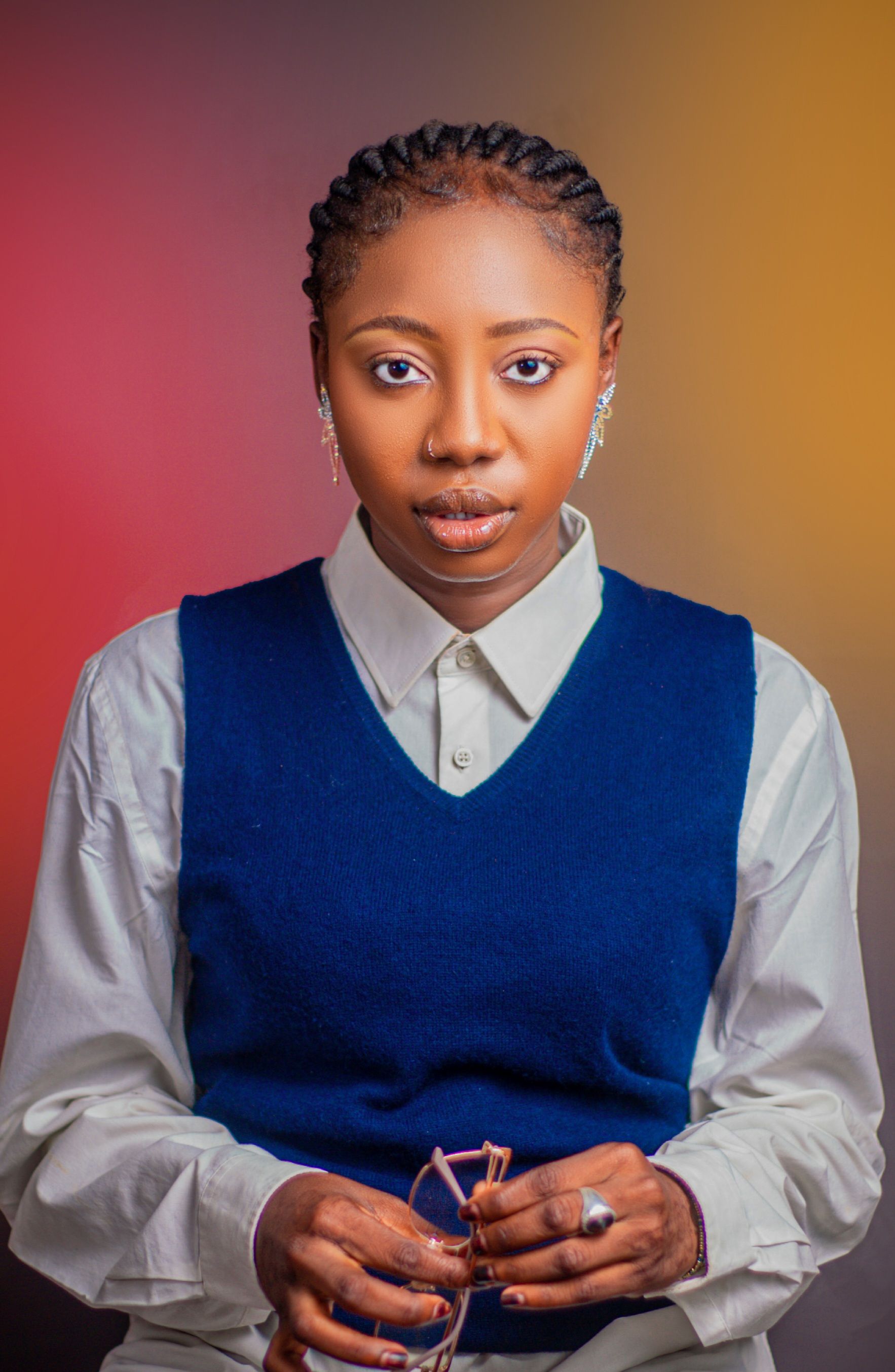 A young woman with braided hair in a stylish blue vest poses thoughtfully.
