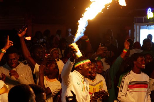 A lively night celebration in Senegal with a man holding a firework as people cheer around him.