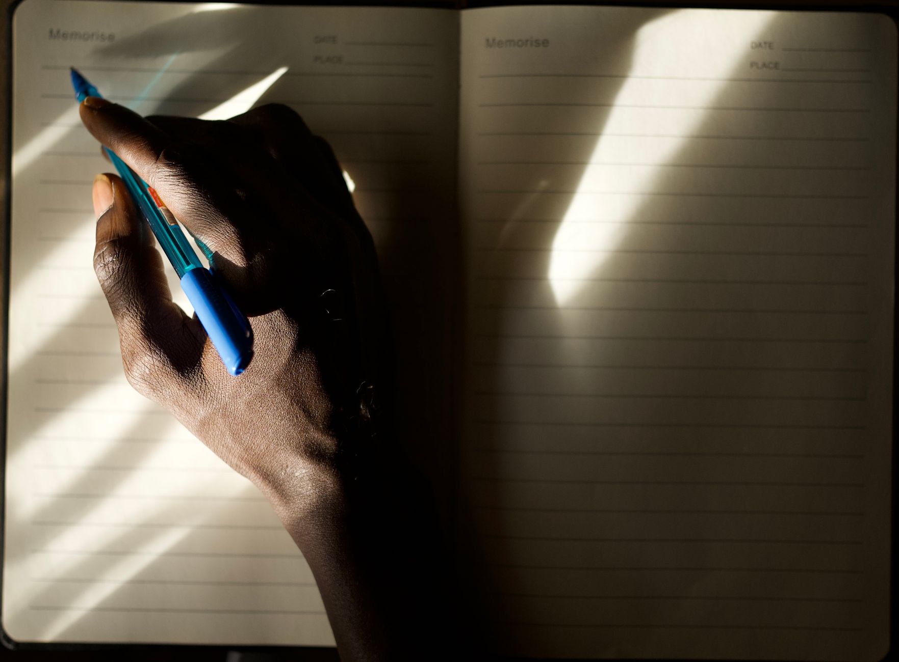 An individual writes notes in a lined notebook, captured in dramatic light and shadow.