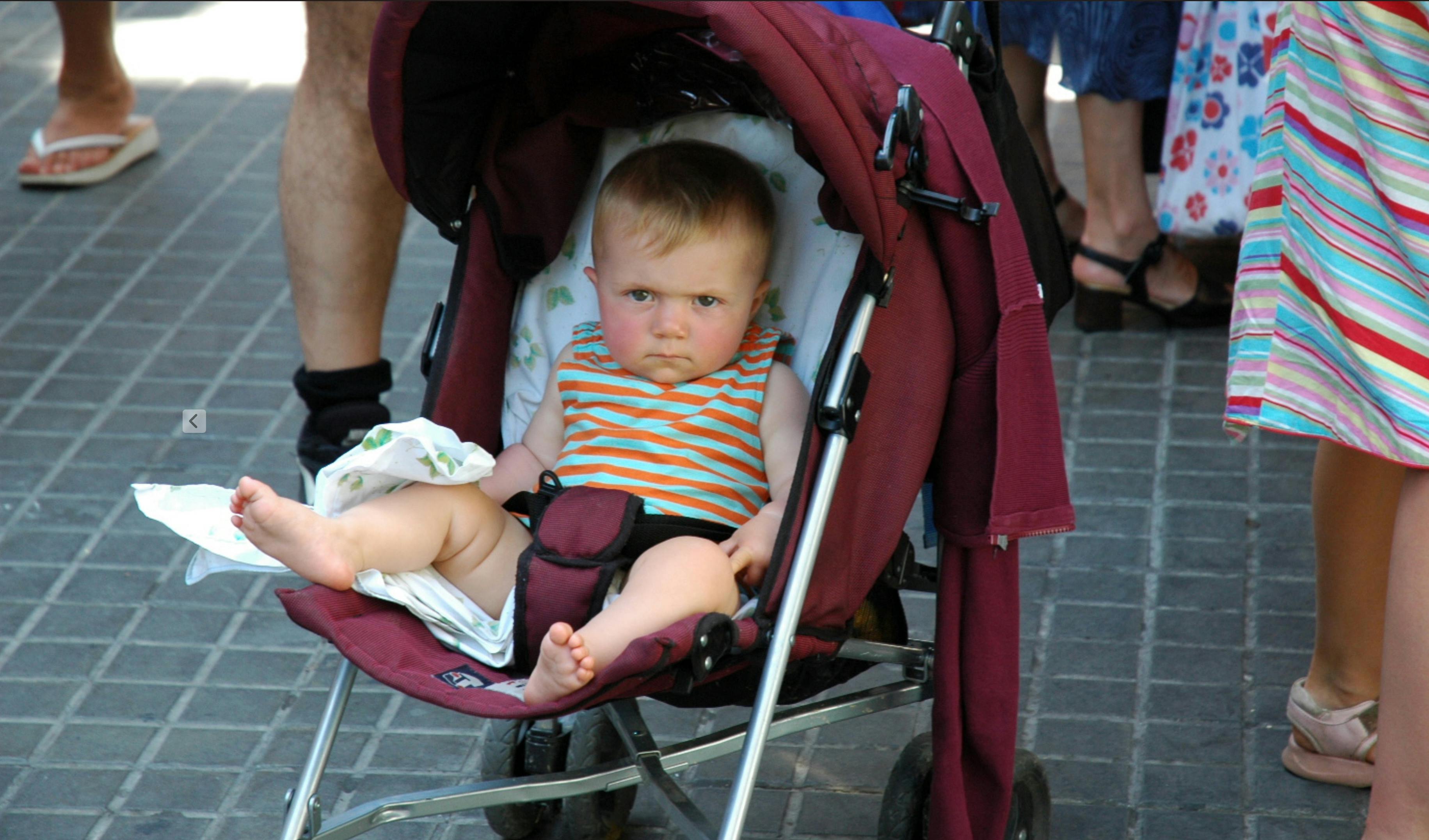 ingyenes Egy baba babakocsiban, komoly arckifejezéssel, emberekkel körülvéve egy napsütéses napon. Stockfotó