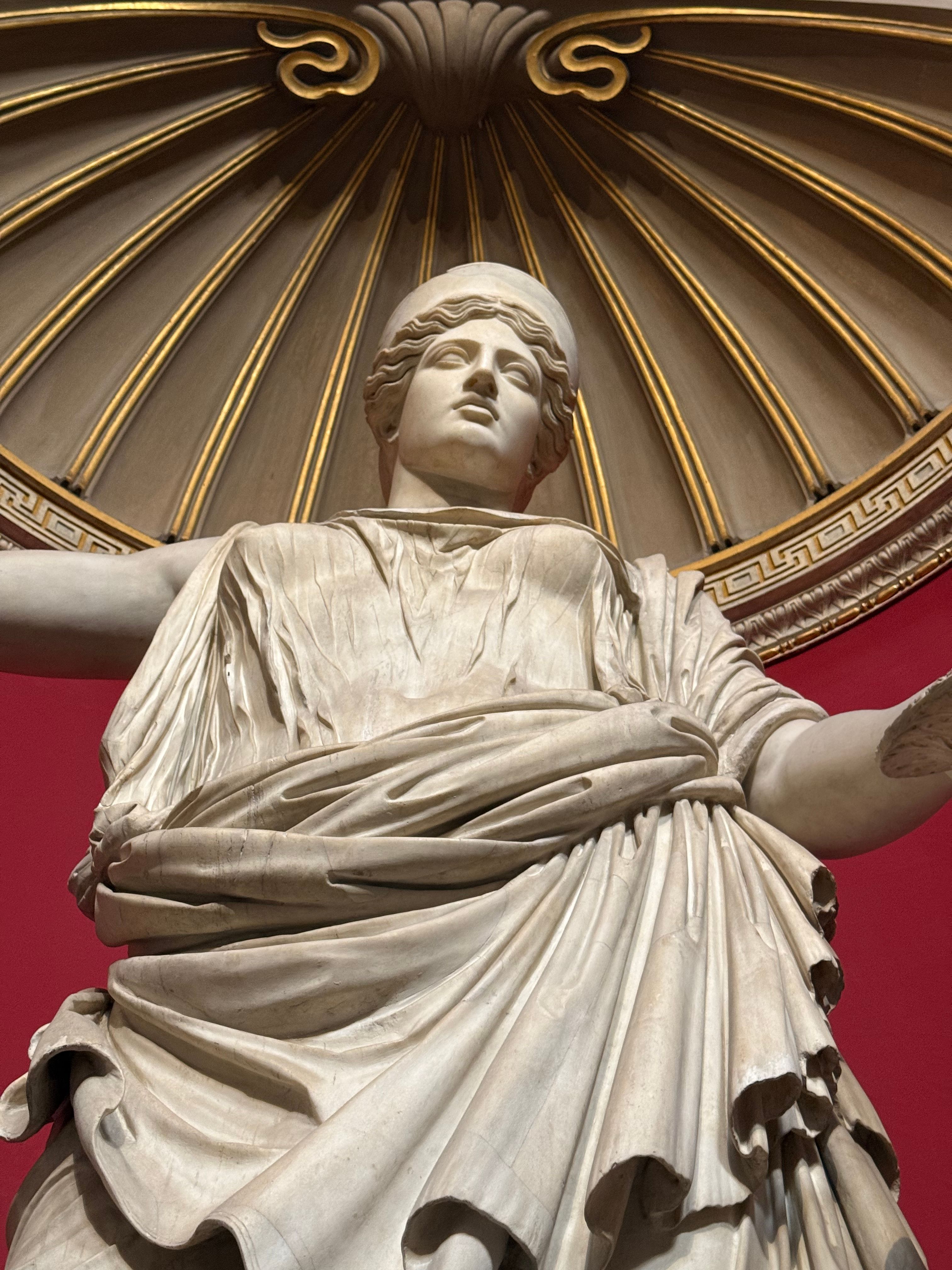 Intricate marble statue with draped garments under ornate ceiling, showcasing classical art elements.