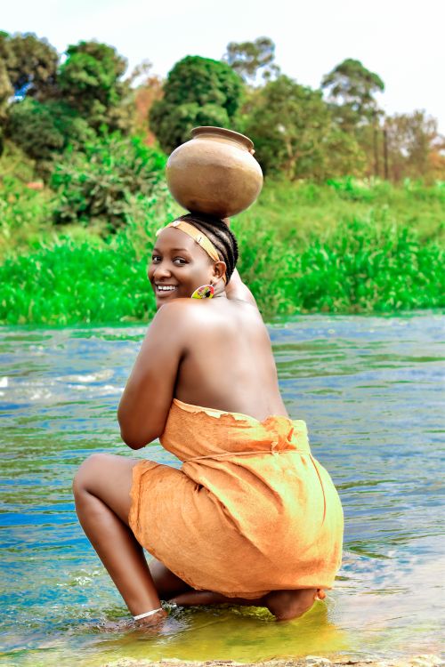 Free A woman squats by a river, carrying a traditional pot on her head in a natural setting. Stock Photo