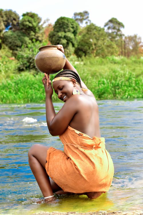Free A woman in traditional attire collecting water by a river, surrounded by lush nature. Stock Photo