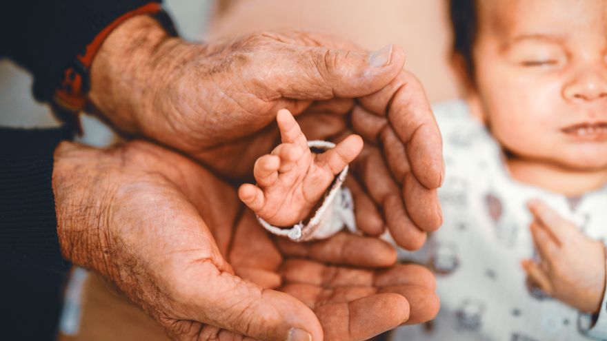 A senior person's hands gently cradling a newborn's hand, representing the inheritance passed down through the male and female lines of a Destiny Matrix chart