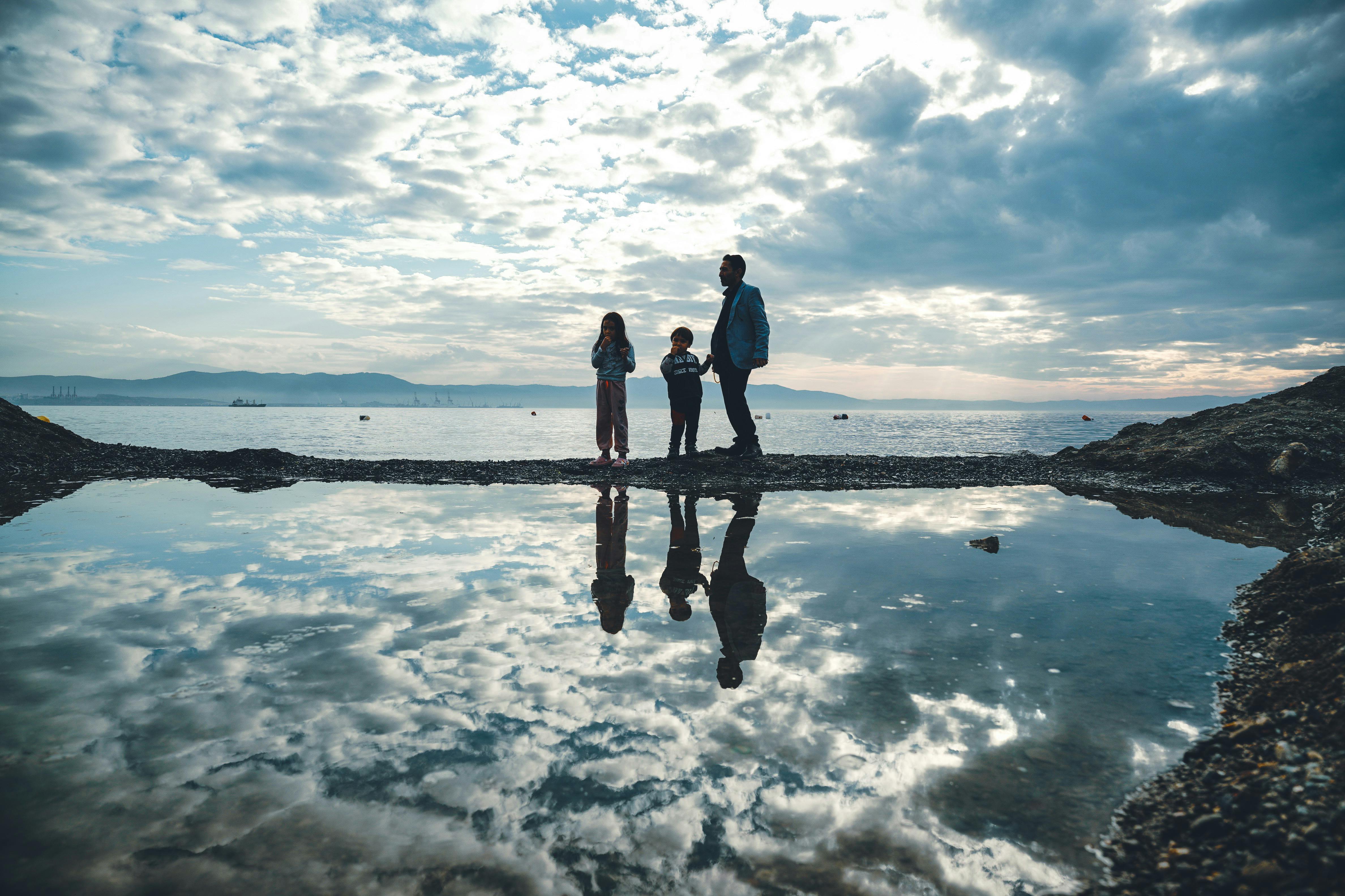 ฟรี ภาพเงาครอบครัวอันสงบสุขริมทะเลในยามพลบค่ำ สะท้อนอยู่ในแอ่งน้ำริมทะเลตัดกับท้องฟ้าที่ปกคลุมไปด้วยเมฆ คลังภาพถ่าย