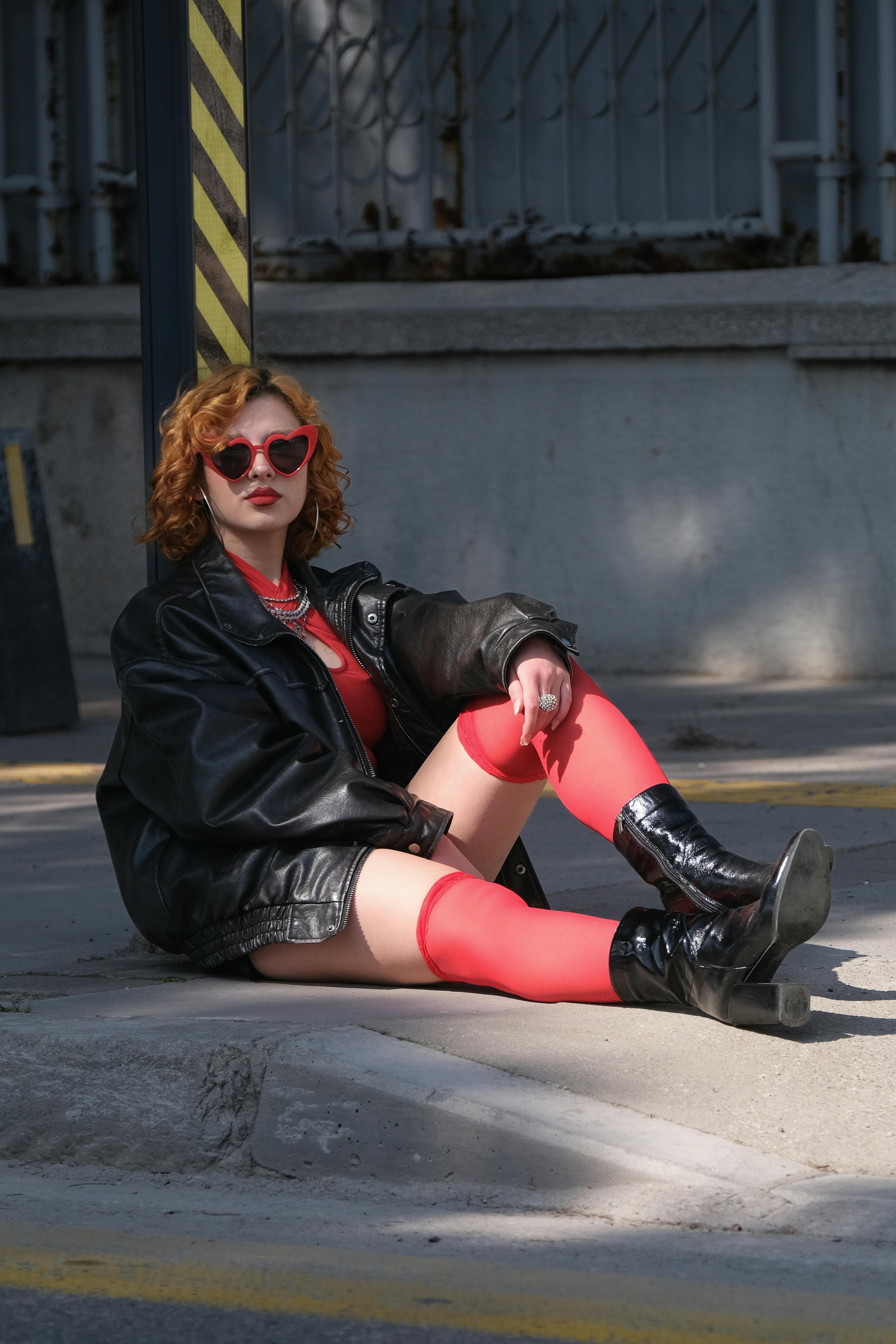 免費 一位穿著紅色緊身褲和黑色皮夾克的時尚女性坐在城市街道上。 圖庫相片