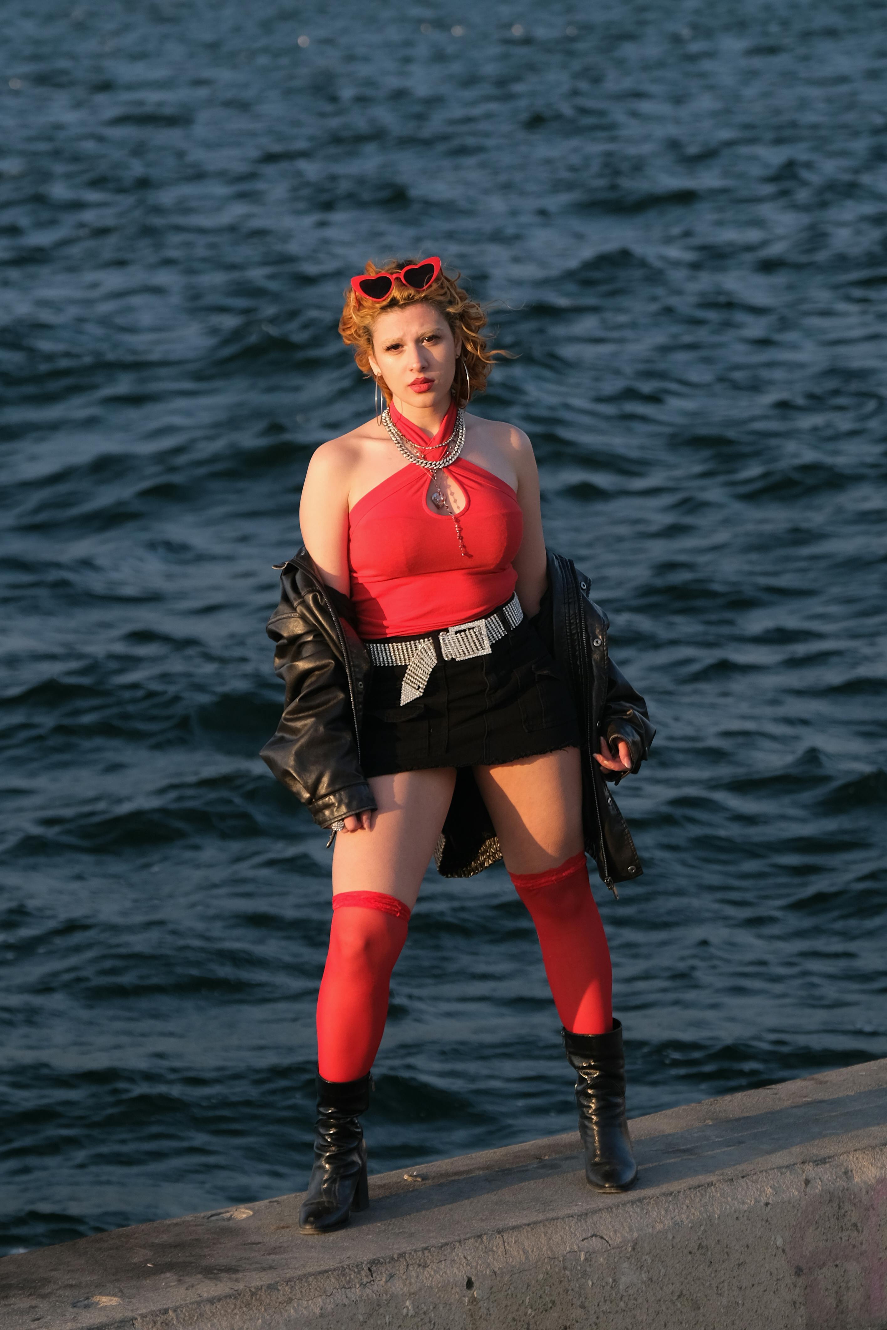 무료 세련된 여성이 빨간색 옷과 가죽 재킷을 입고 낮에 물가에서 포즈를 취하고 있다. 스톡 사진