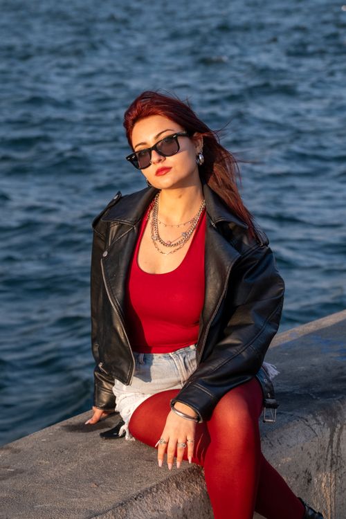 Free A fashionable woman in sunglasses and a leather jacket sits near the water's edge during the day. Stock Photo
