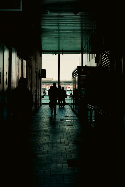 Free Silhouetted figures walk through a dark urban alleyway leading to a distant outdoor view. Stock Photo