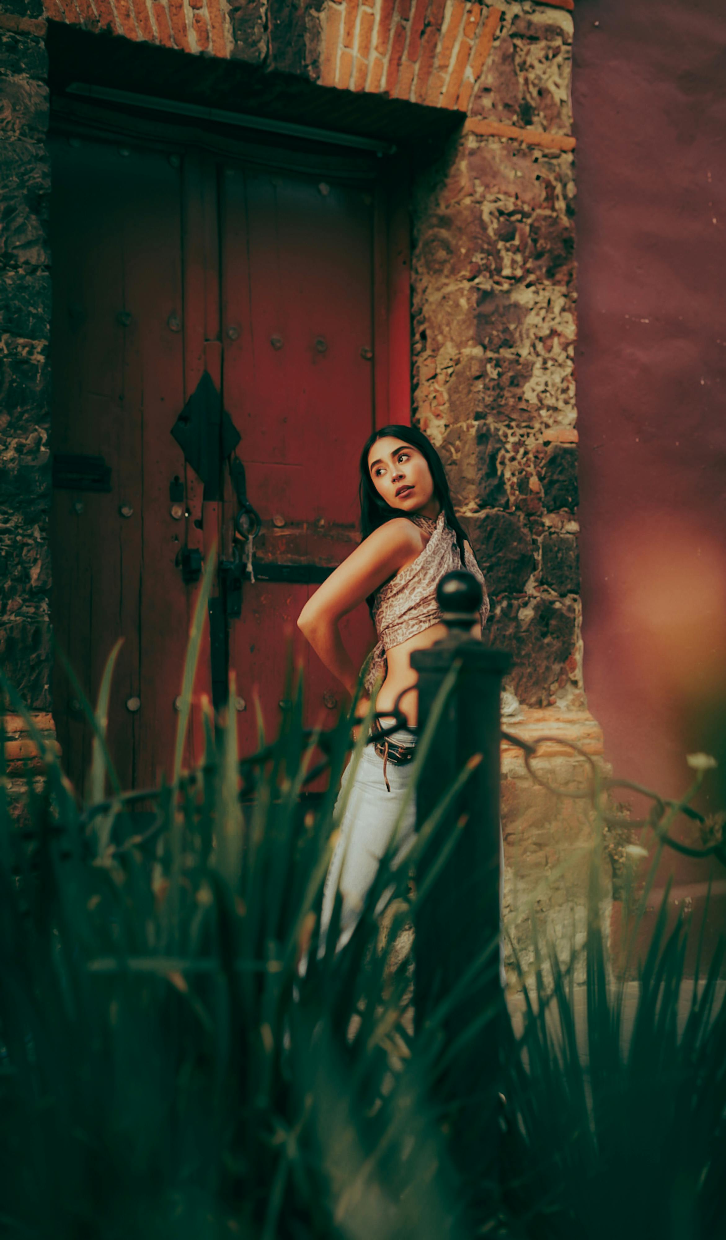 bezplatná Mladá žena elegantně pózuje u rustikálních červených dveří s kamennými akcenty v městském prostředí. Základová fotografie