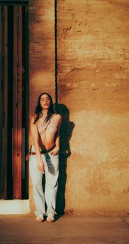 Portrait of a young woman in casual attire standing against a sunlit wall with dramatic shadows.
