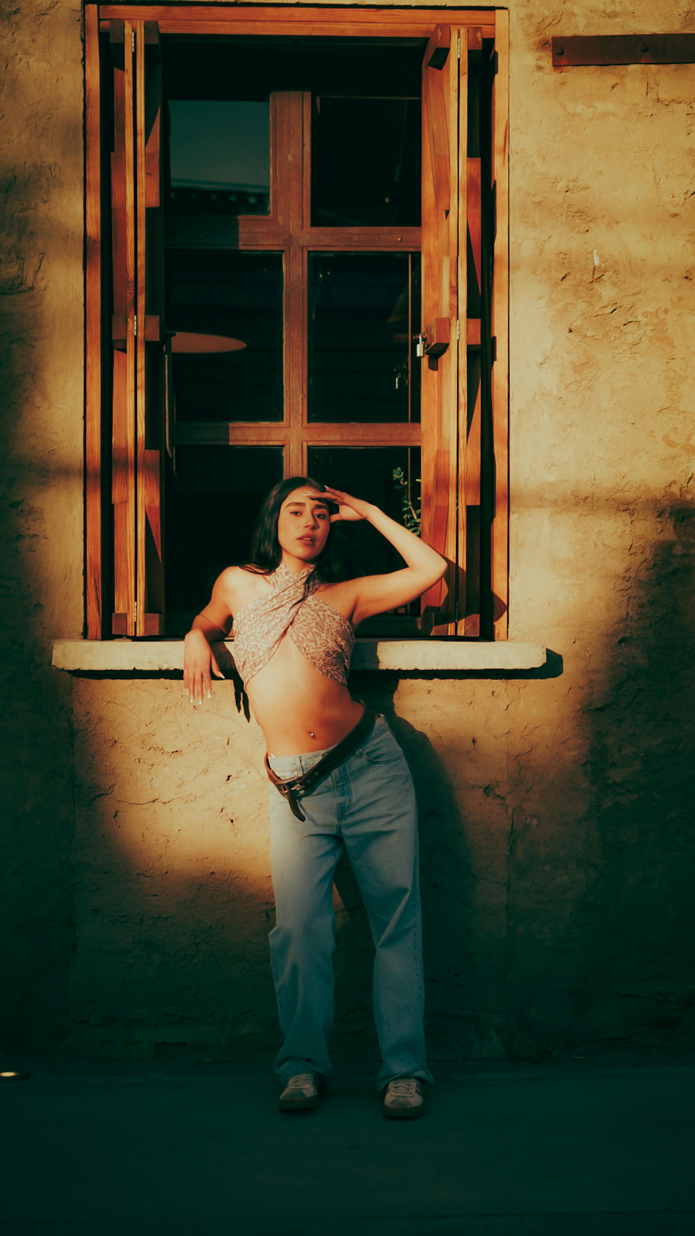 Kostenlos Eine junge Frau steht elegant an einem rustikalen Fenster, das von warmem Sonnenlicht erhellt wird und ein kunstvolles Schattenspiel erzeugt. Stock-Foto