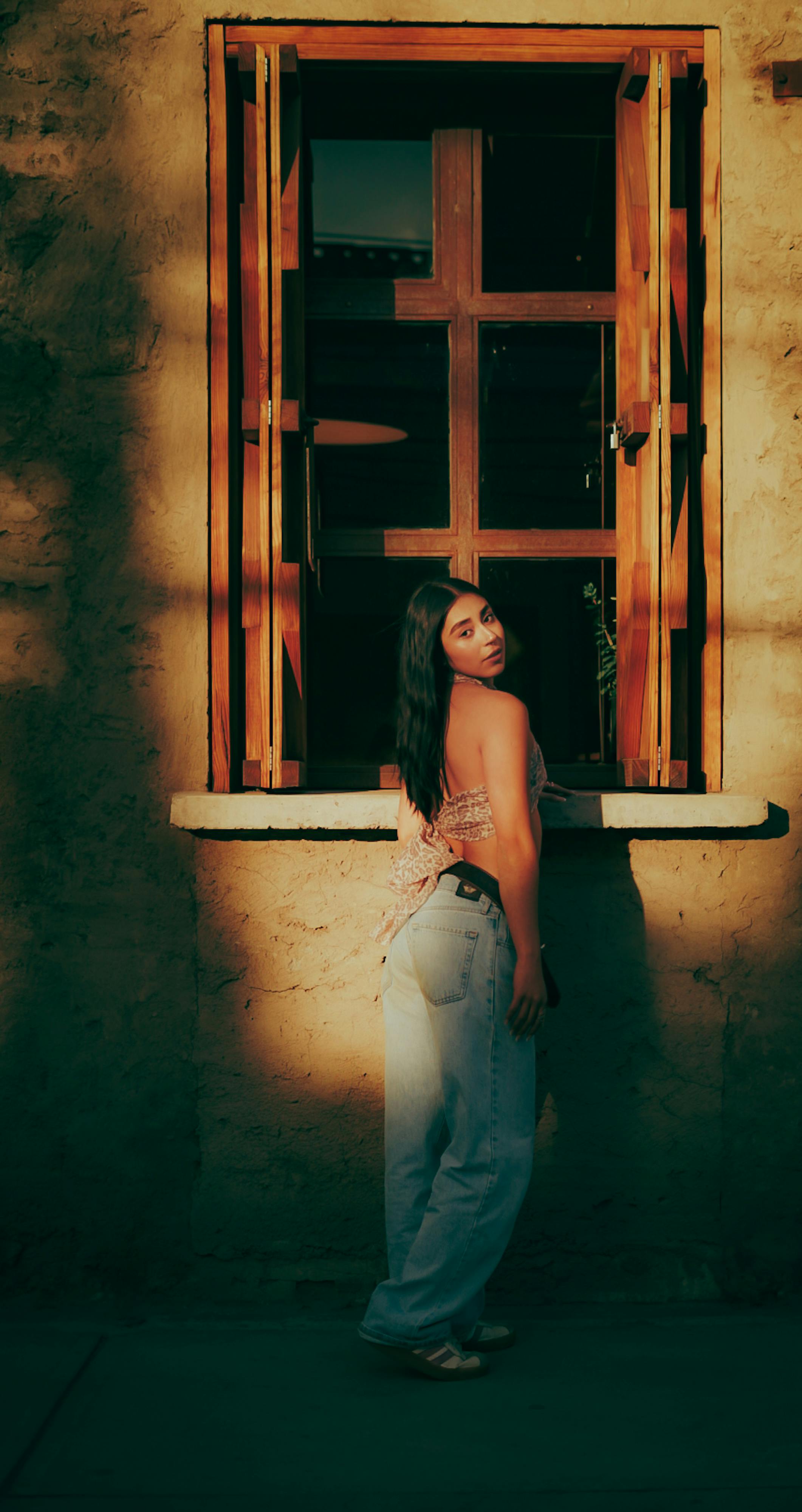 Zadarmo Štýlová žena v ležérnom oblečení pózuje pri slnkom zaliatom okne, ktoré vrhá teplé tiene. Fotka z fotobanky