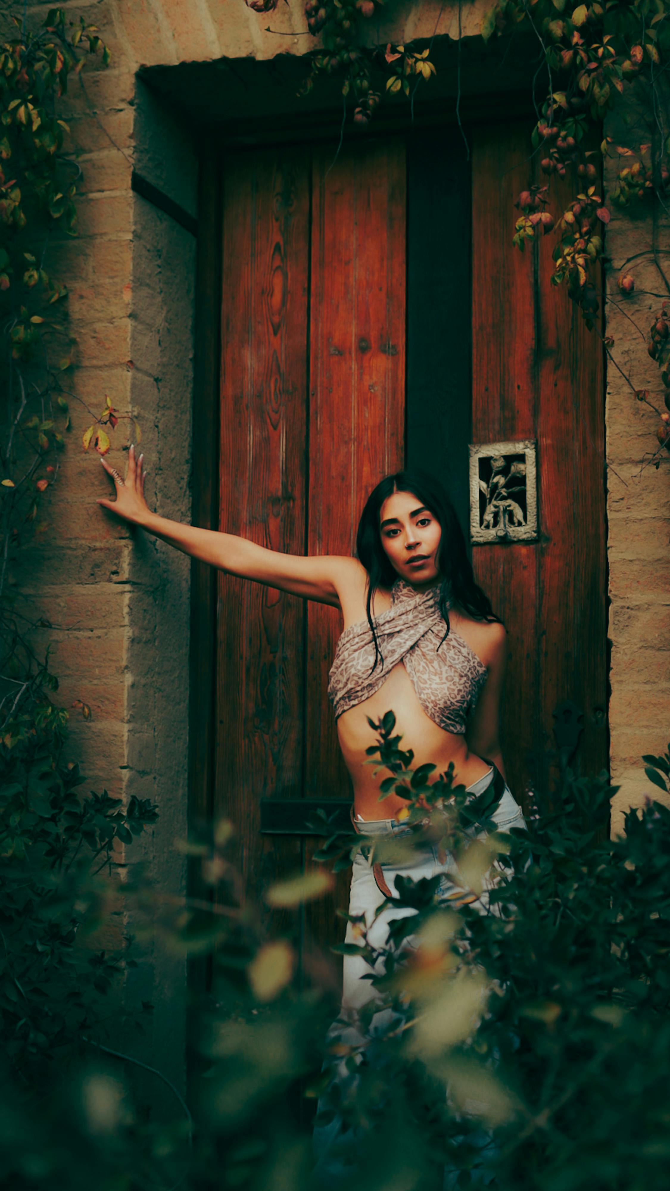 ฟรี หญิงสาวทันสมัยในชุดมีสไตล์โพสท่าอยู่ข้างประตูไม้สไตล์ชนบทที่ล้อมรอบด้วยใบไม้ คลังภาพถ่าย