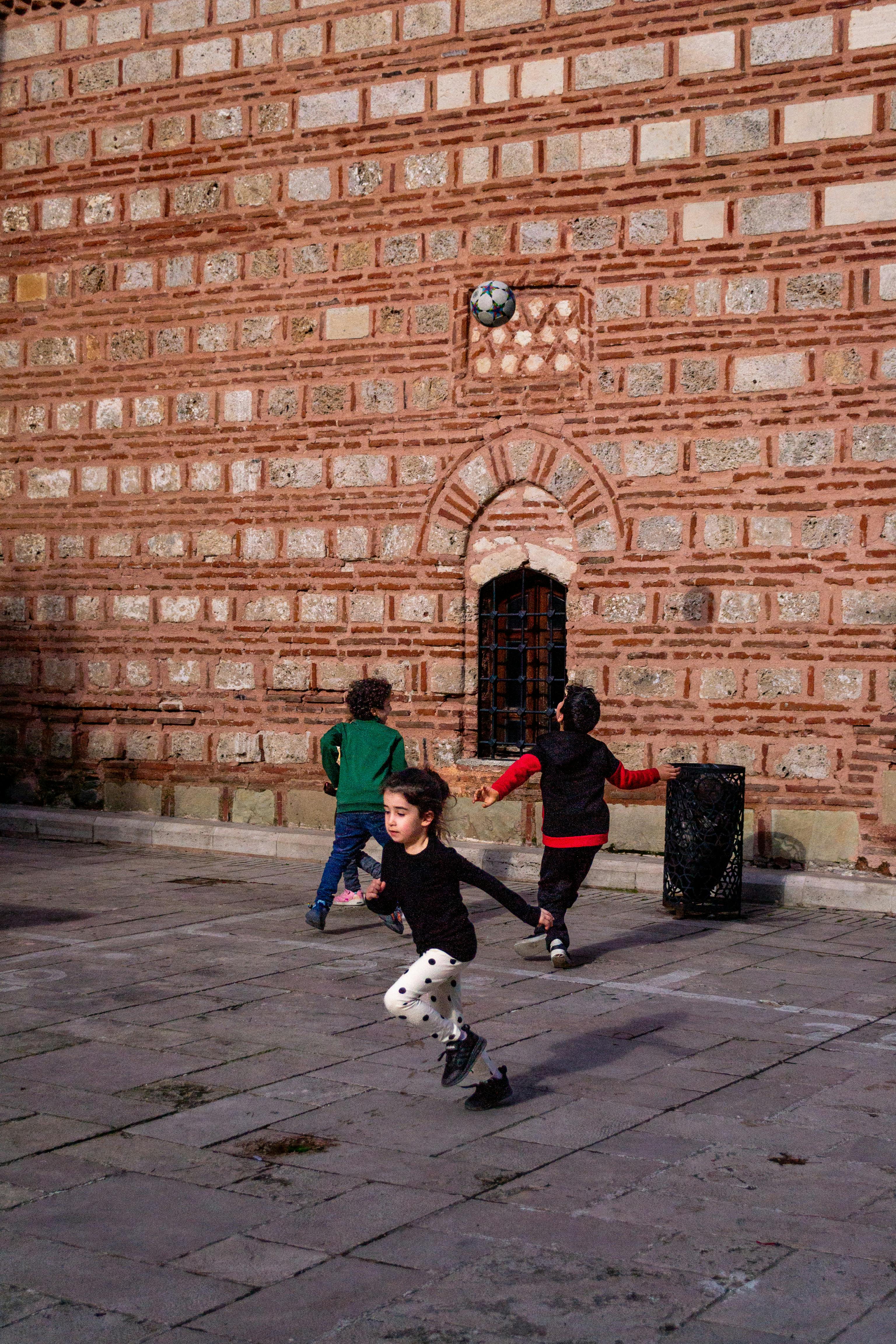 Безкоштовне Група дітей грає у футбол у внутрішньому дворику з історичною цегляною стіною на тлі. стокове фото