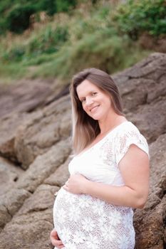 A joyful pregnant woman in a white dress smiles near rocky terrain. Perfect for parenting and maternity themes.