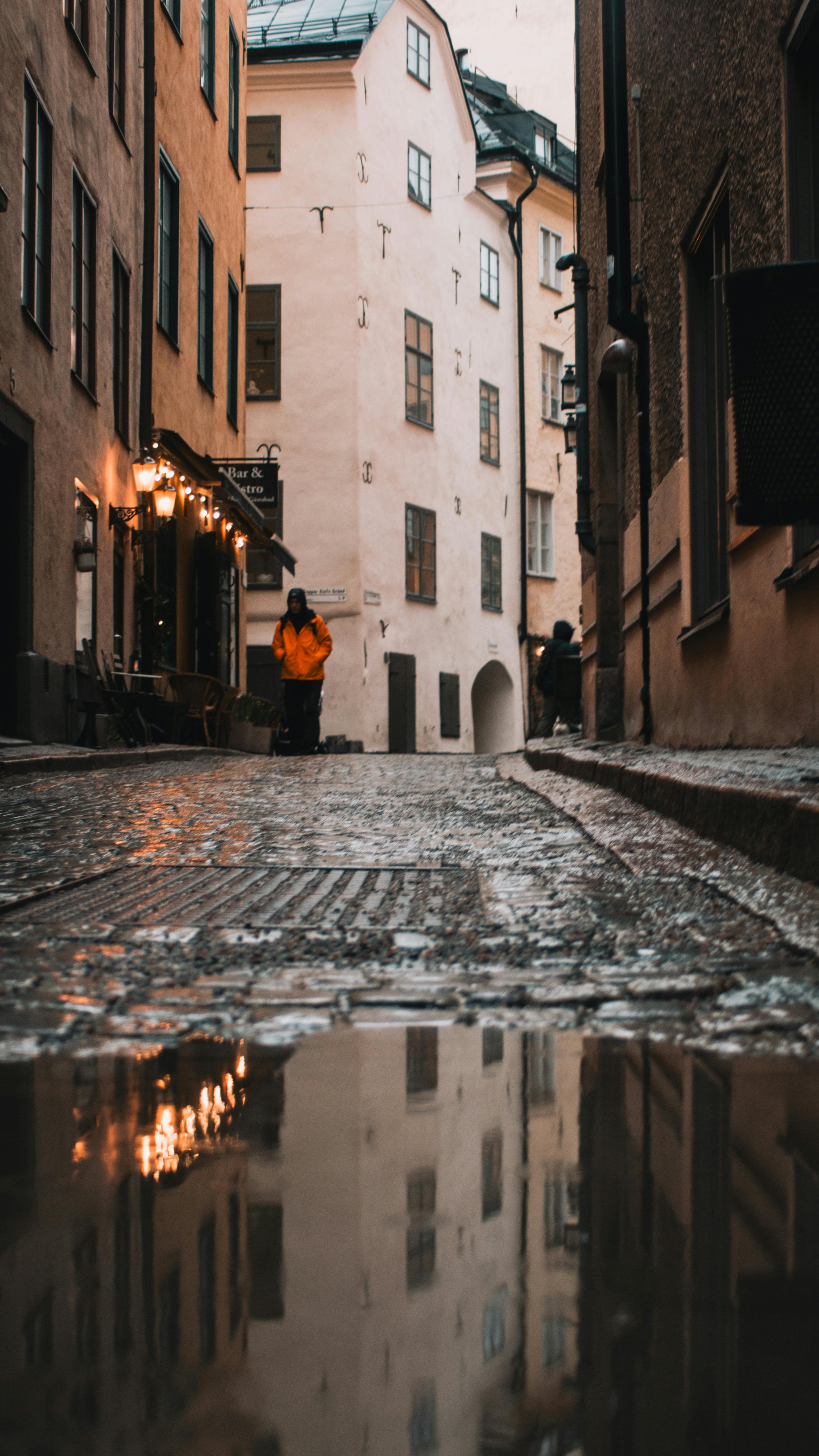 Безкоштовне Тепло освітлений провулок у Стокгольмі створює затишну осінню атмосферу з відображеннями в калюжі, вловлюючи безтурботний міський шарм. стокове фото