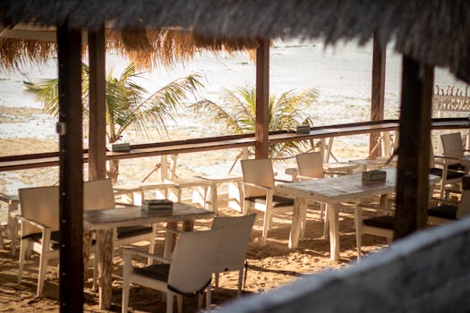 Serene beachside restaurant with empty tables and chairs, under thatched roof, overlooking palm trees and sand.