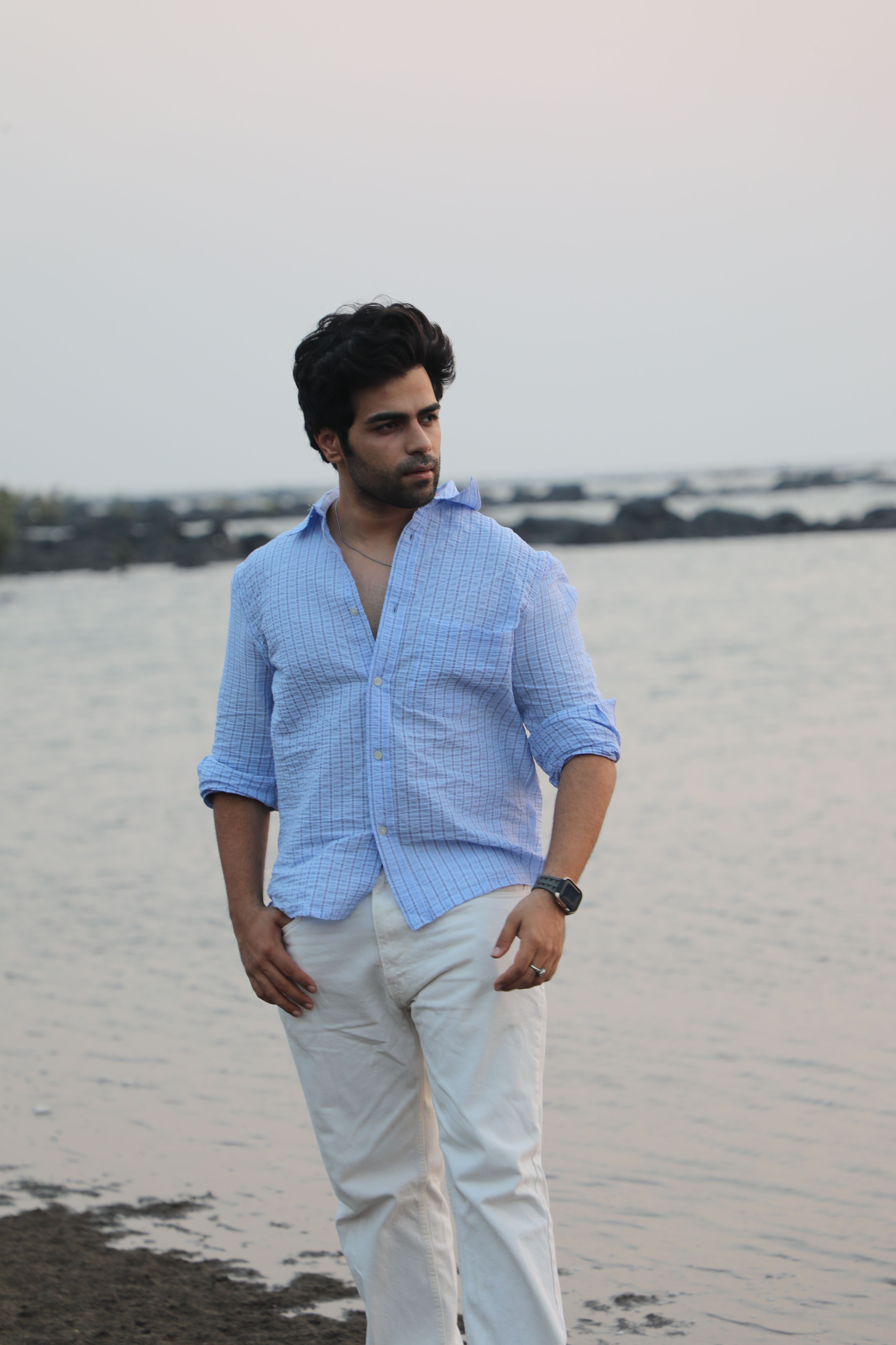 Young man in blue shirt posing by the sea at sunset, exuding a relaxed vibe.