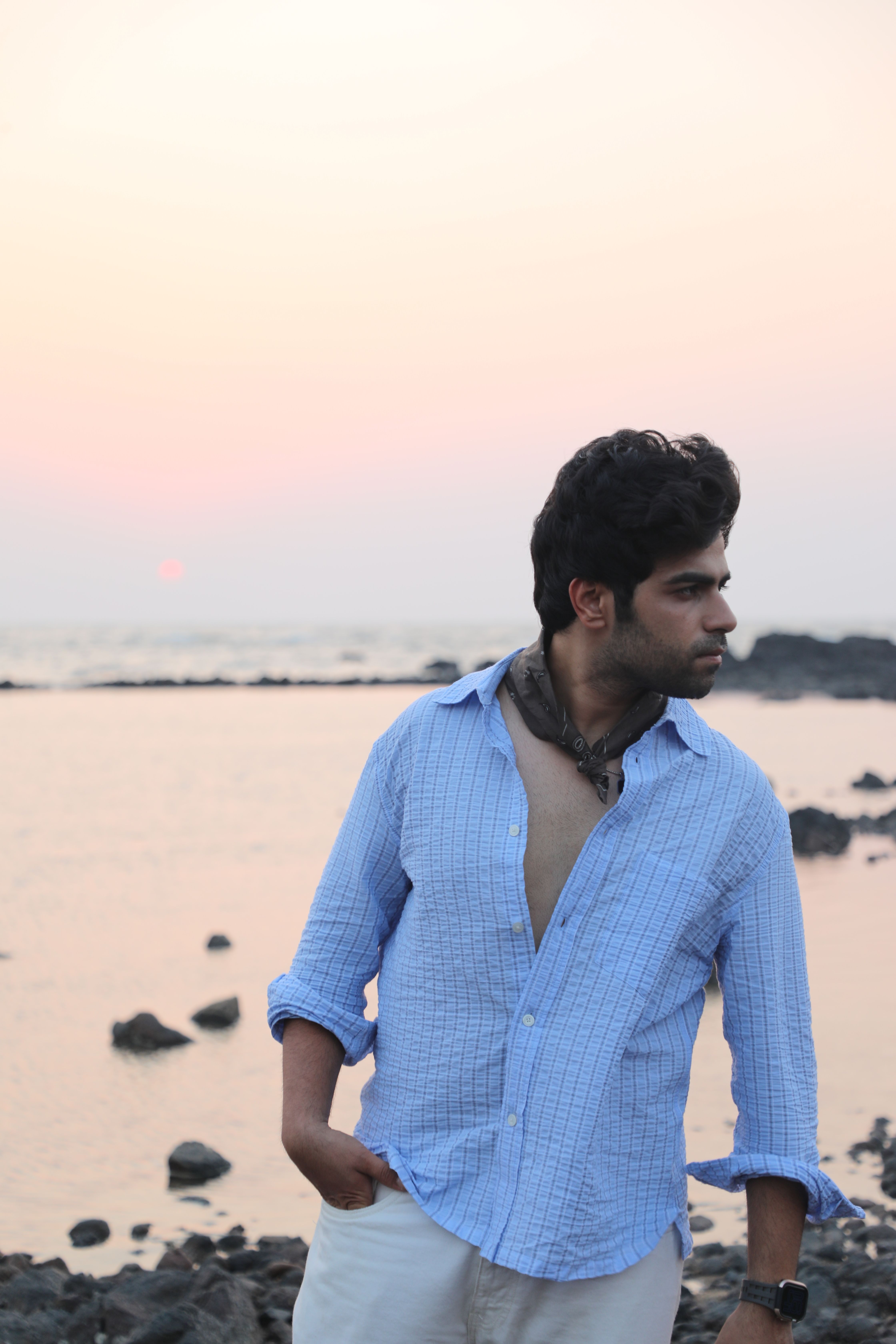 Young man in a blue shirt standing on a rocky beach at sunset, gazing into the distance.