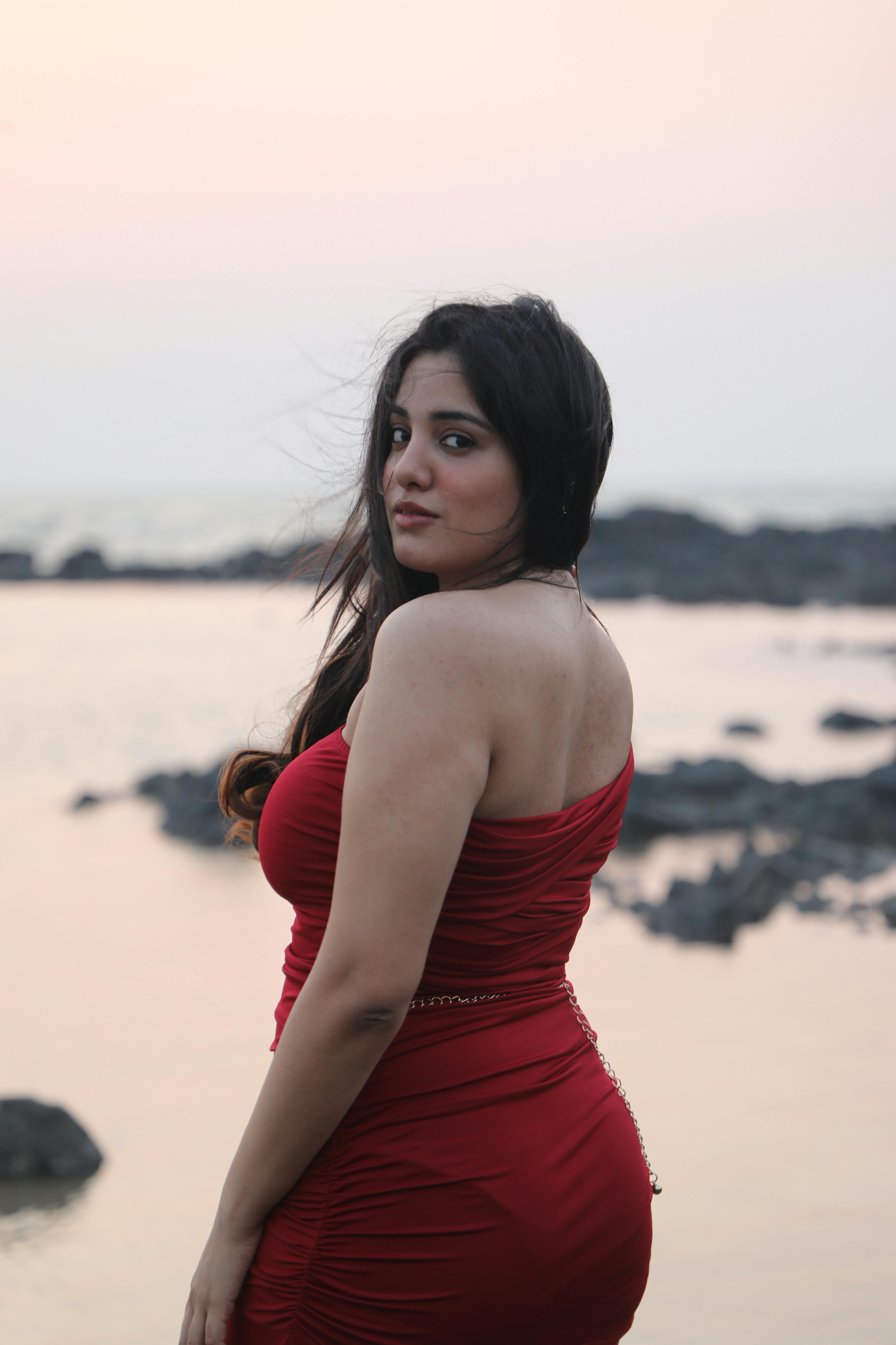 Woman in a red dress posing by the rocky seashore at sunset, exuding elegance and grace.