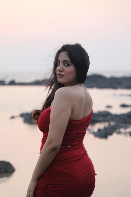 Woman in a red dress posing by the rocky seashore at sunset, exuding elegance and grace.