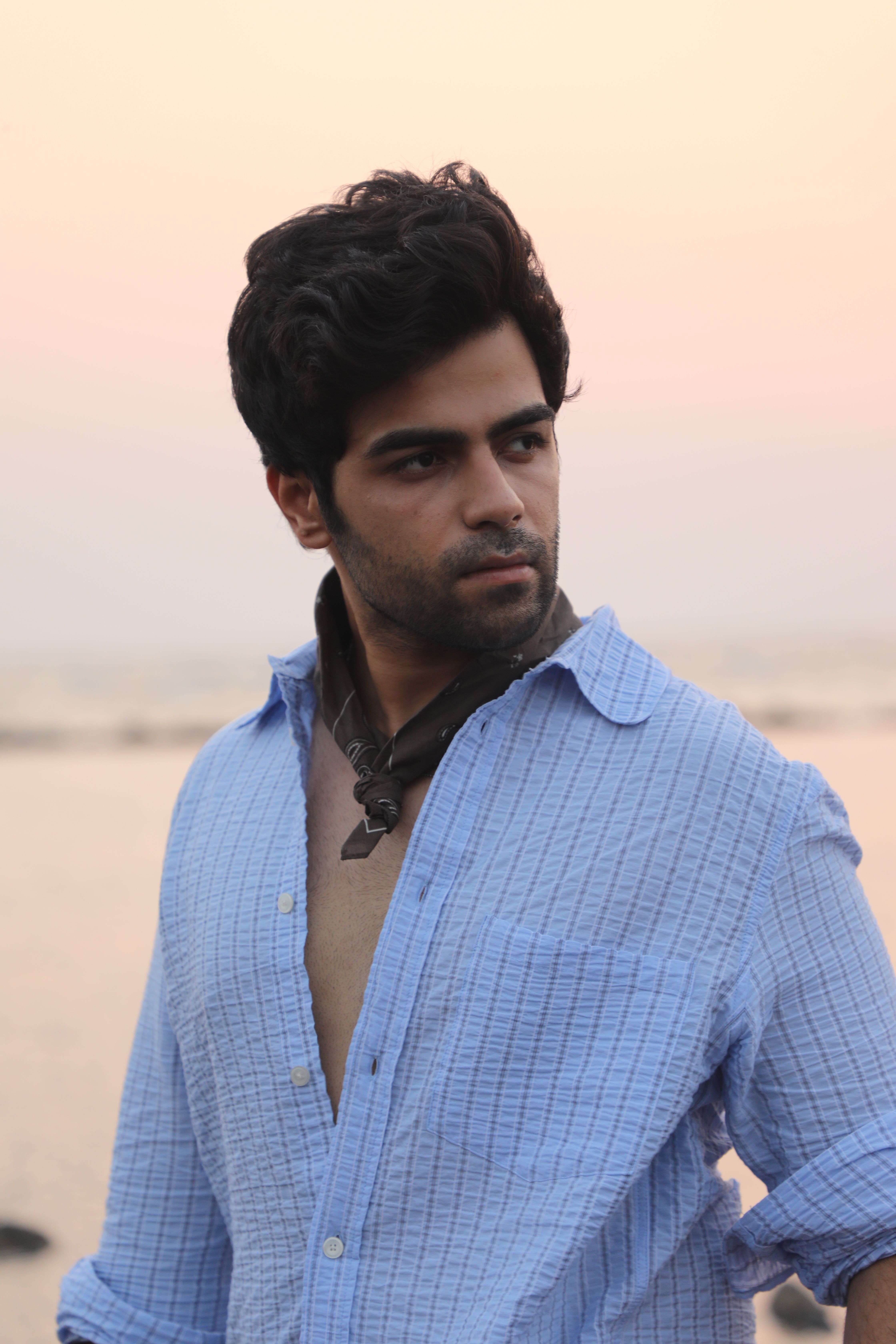 Stylish young man posing outdoors with a sunset beach background, wearing a casual blue shirt.