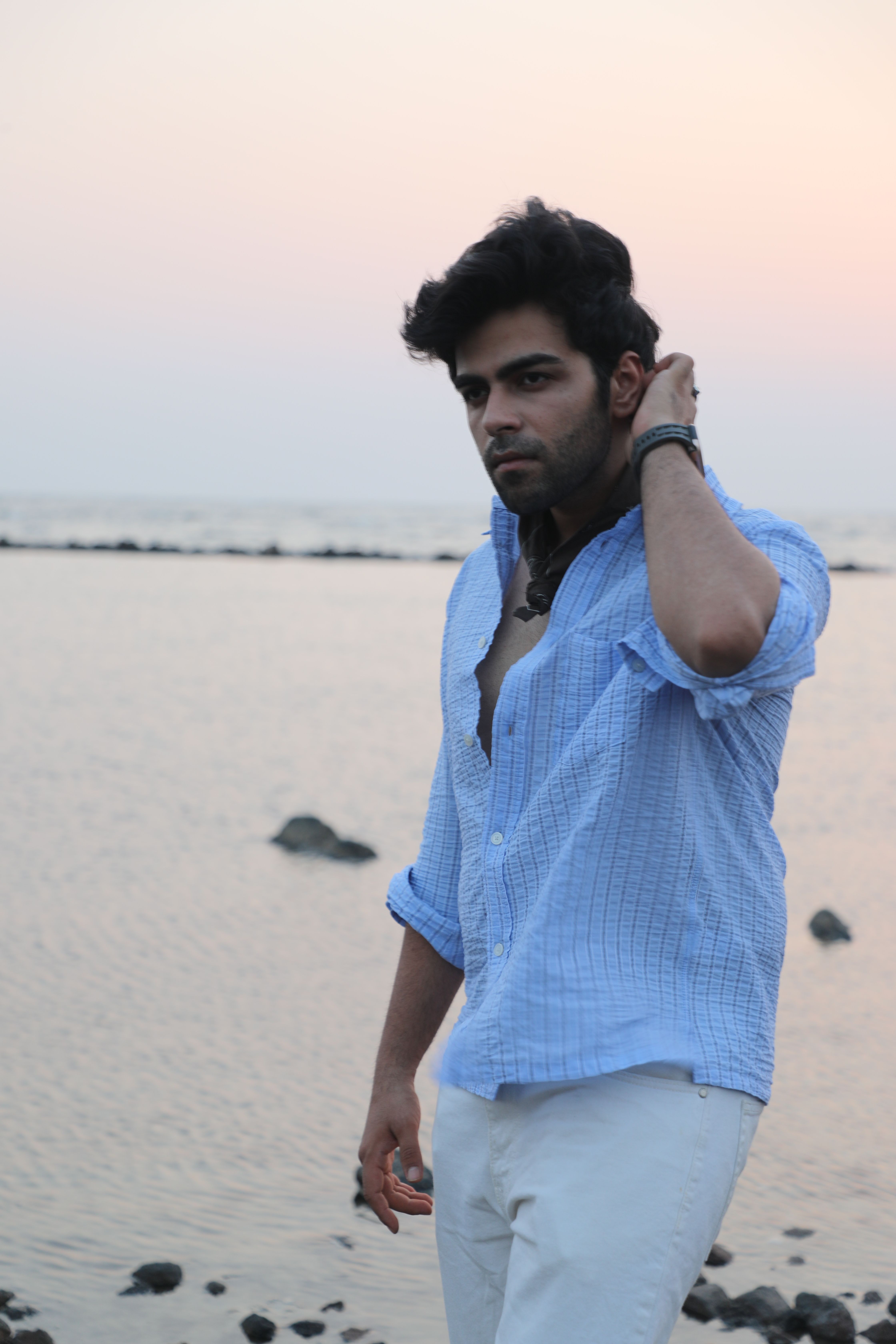 A man in a blue shirt poses by the sea at sunset, exuding confidence and style.
