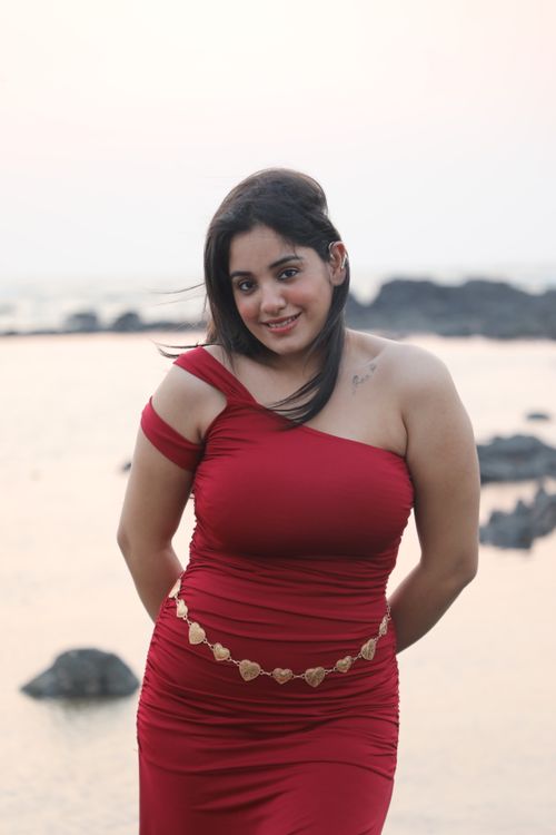 Free A woman in a red dress poses by the ocean at sunset, exuding confidence and elegance. Stock Photo