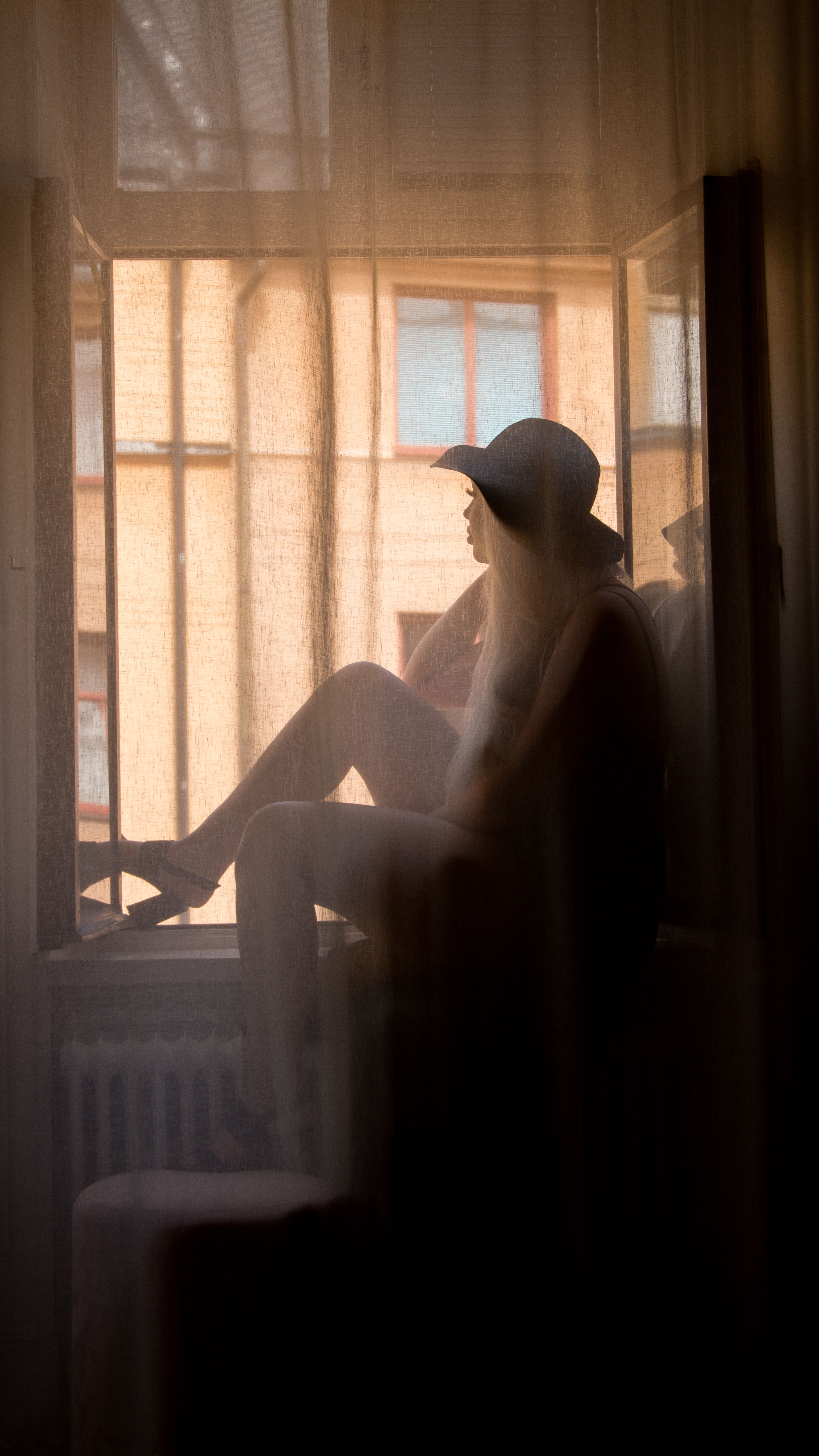 A woman in silhouette sits by an open window wearing a hat in a summer setting.