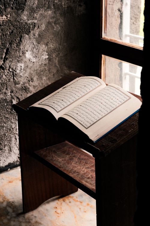 Free A book opened on a wooden stand near a window, illuminated by sunlight in a textured room corner. Stock Photo