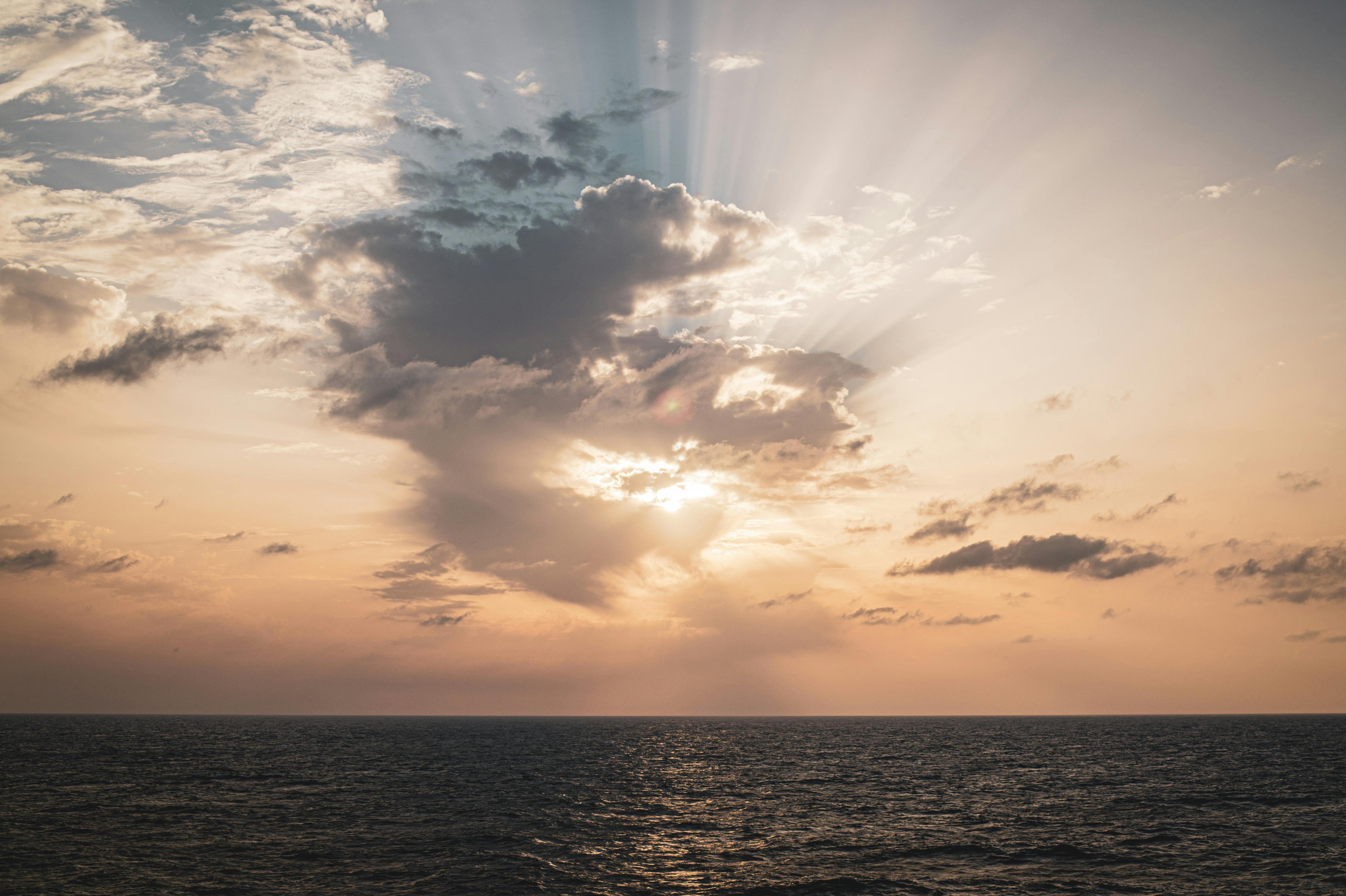 bezplatná Ohromující západ slunce nad oceánem s dramatickými mraky a slunečními paprsky, které pronikají skrz ně a vytvářejí klidnou atmosféru. Základová fotografie