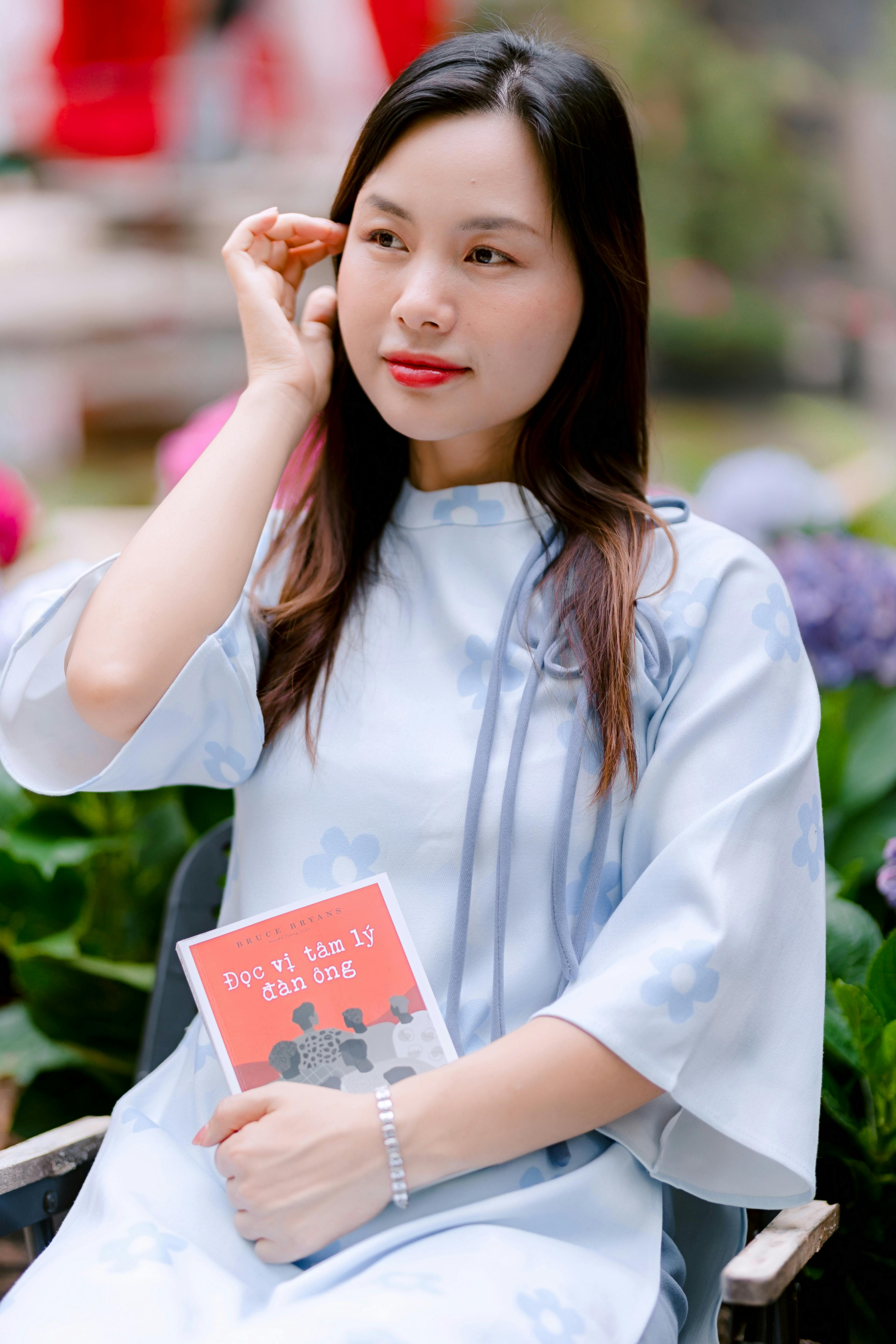 bezplatná Vietnamská žena v tradičním oděvu držící knihu v ruce, sedíc v zahradě. Elegantní a klidná. Základová fotografie