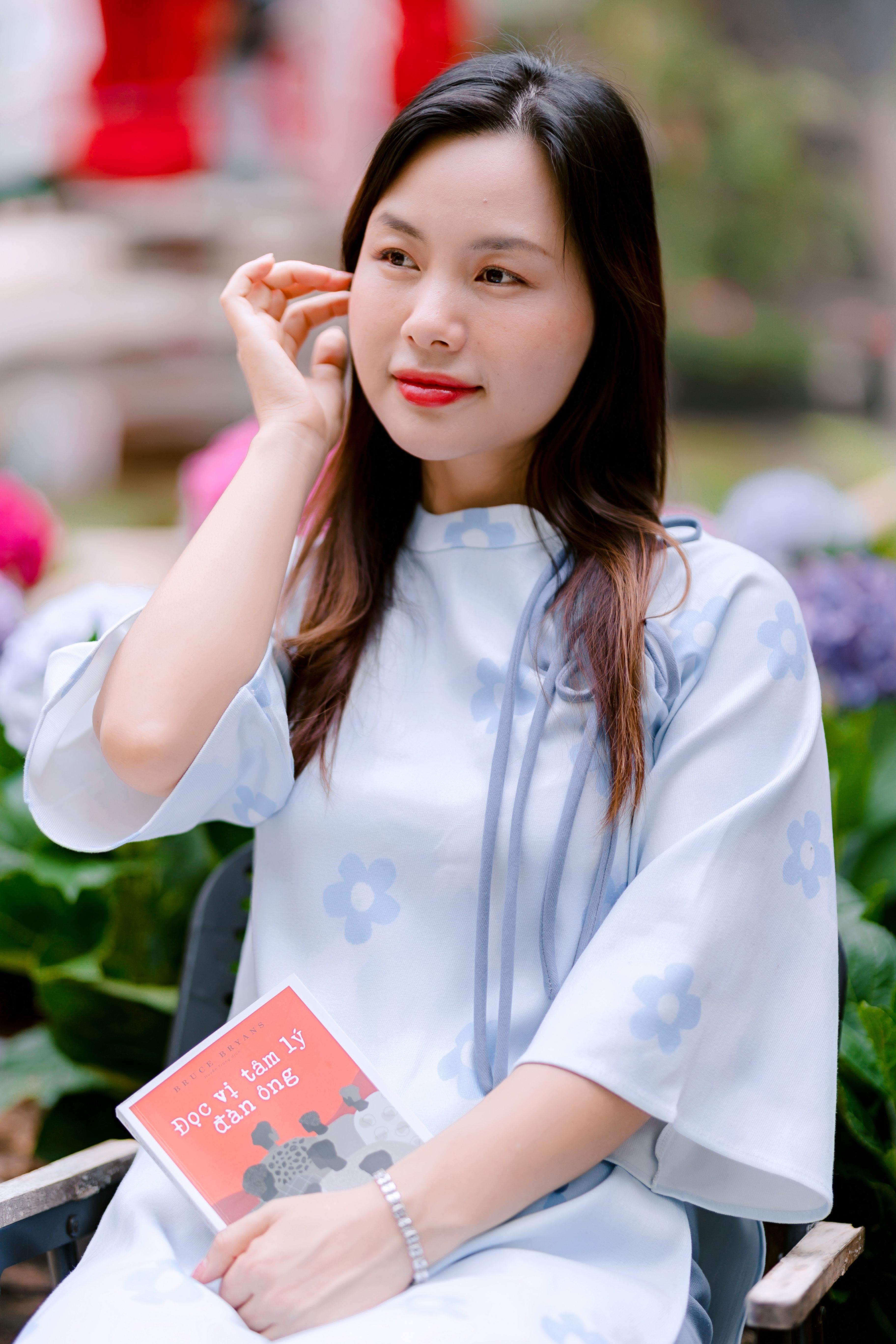 Charming woman in a floral dress enjoys a book while seated outdoors surrounded by flowers.