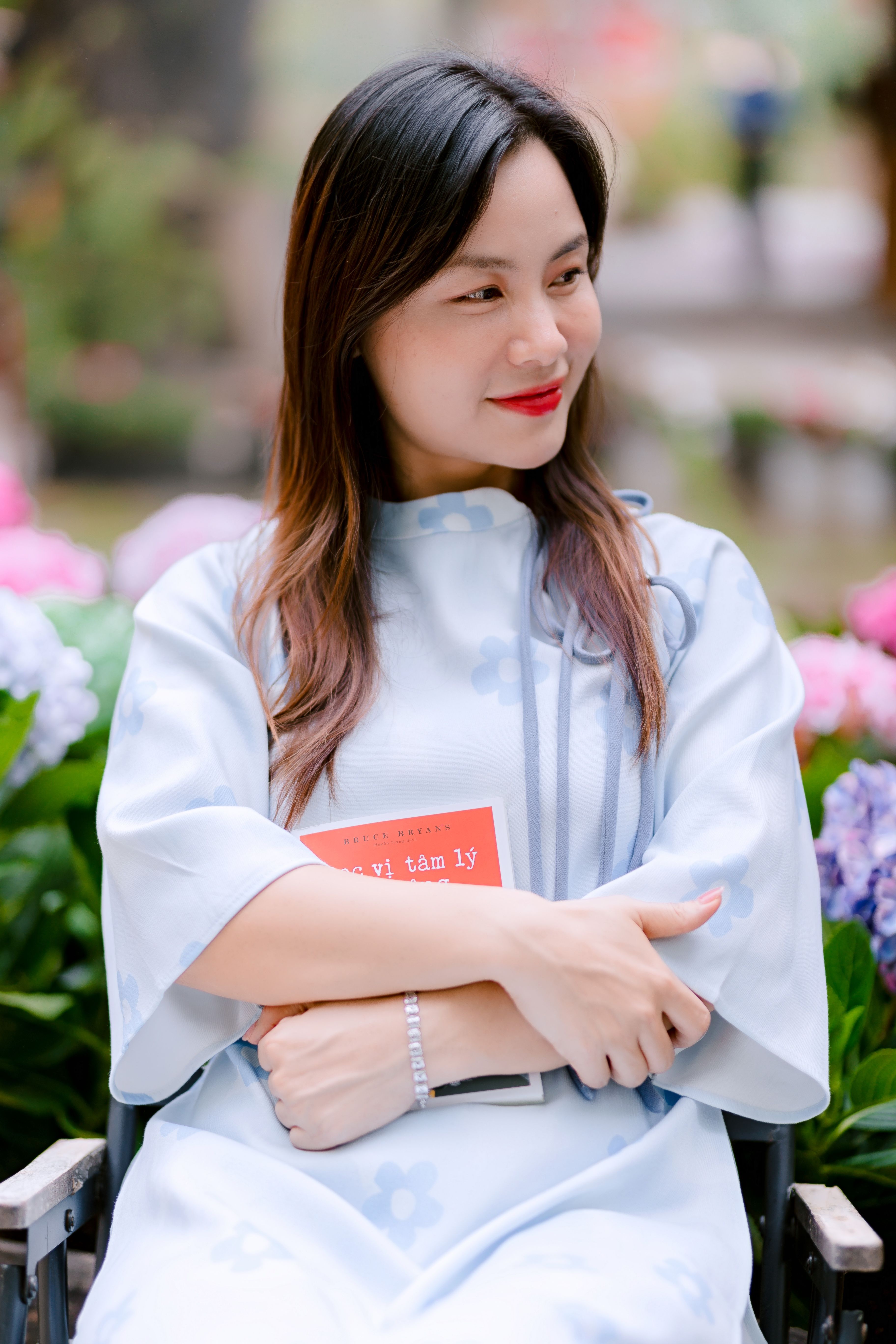 Smiling woman sitting in a garden, holding a book surrounded by colorful flowers.