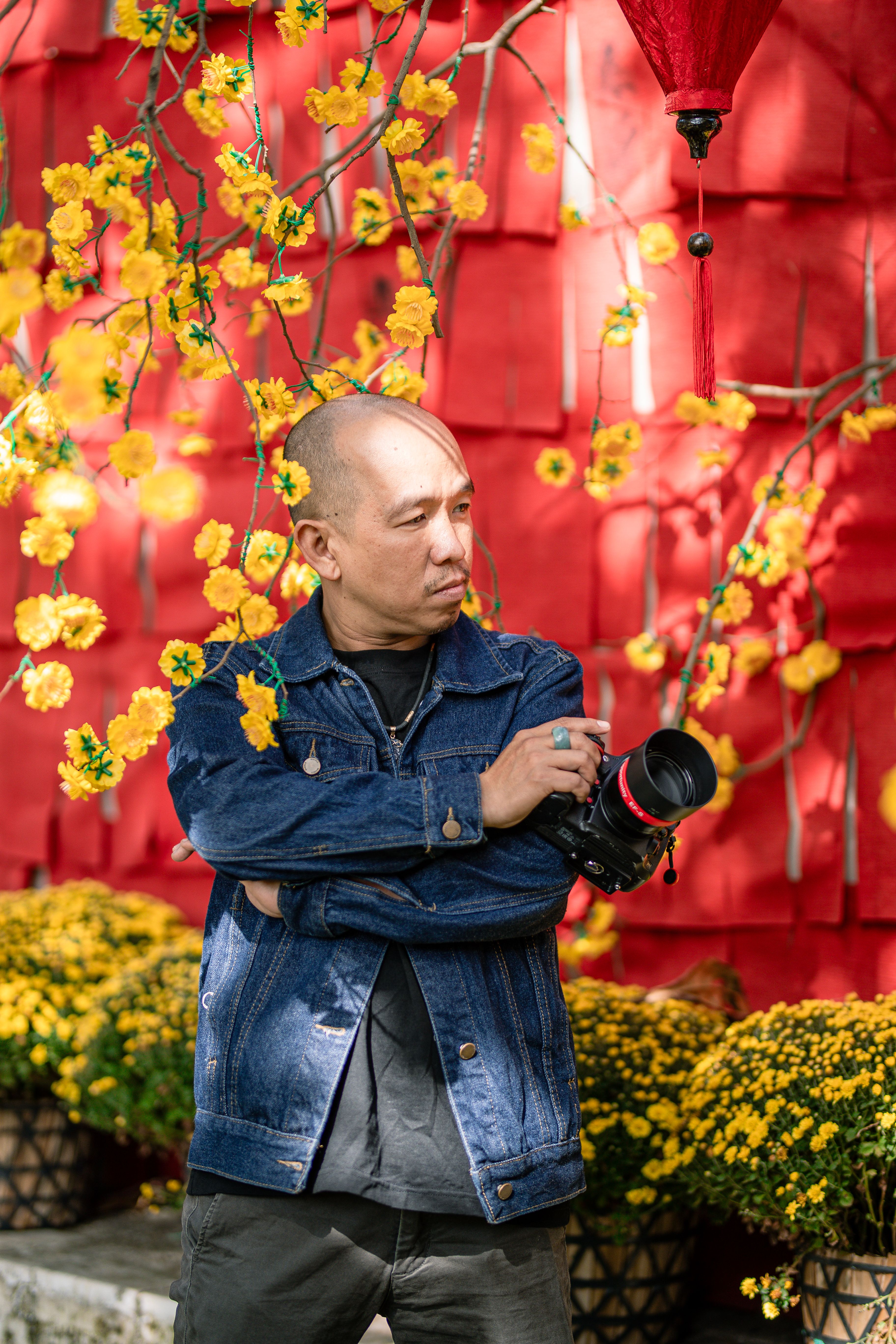 Portrait of a photographer holding camera amidst yellow flowers and red lanterns.