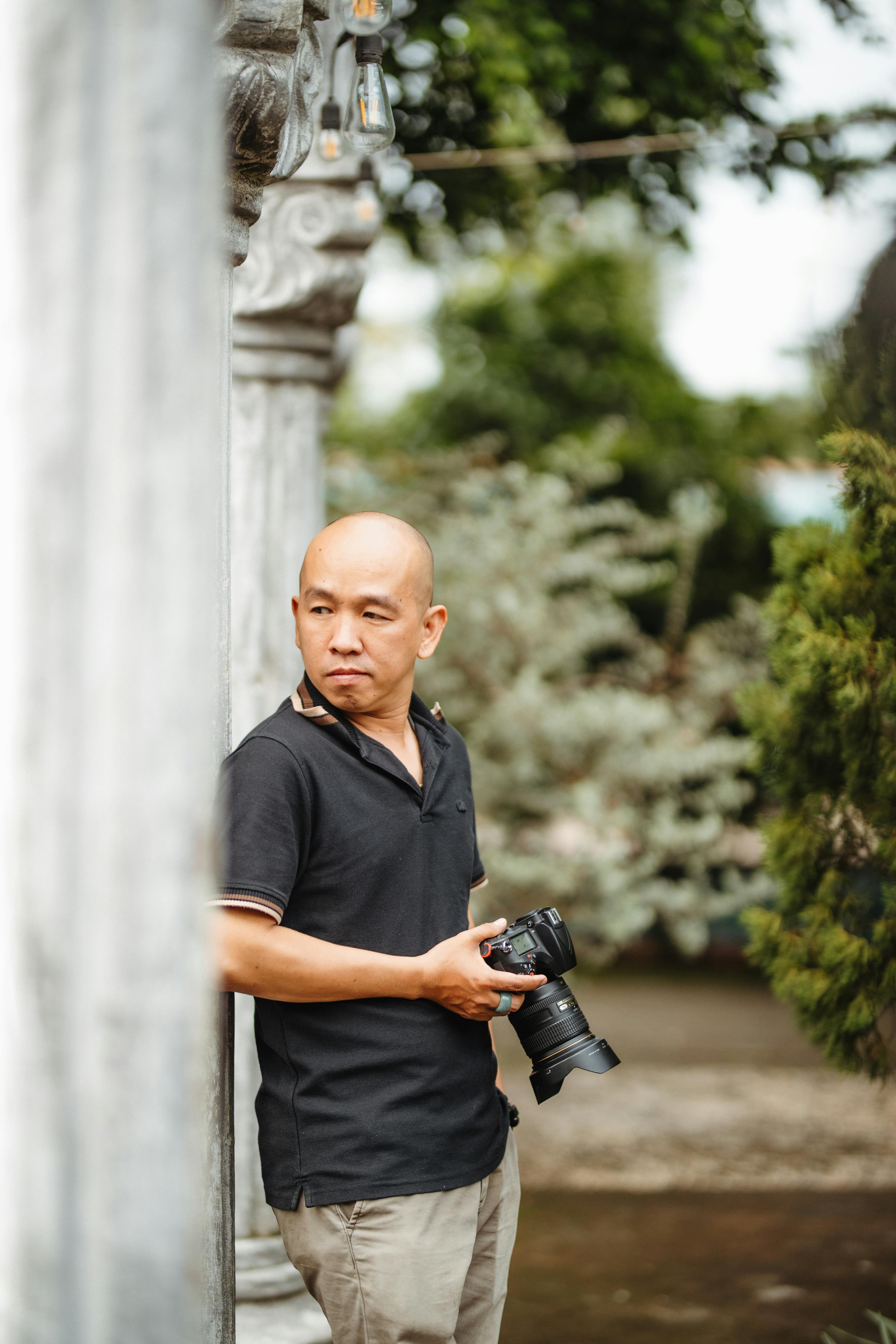 A man standing outdoors with a camera, surrounded by greenery and architectural elements.